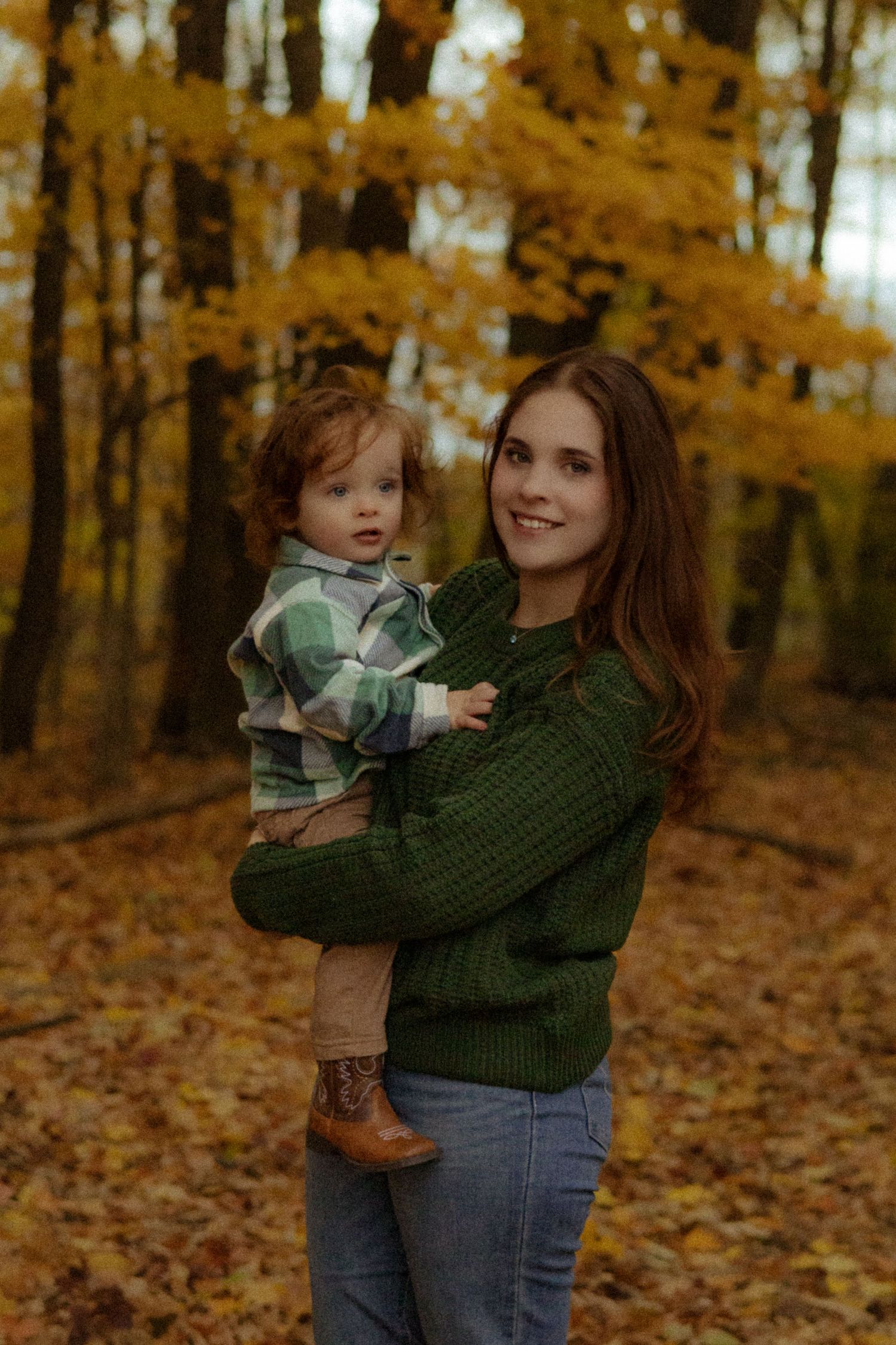 A smiling woman in a green sweater holds a toddler in a plaid shirt amid golden autumn forest leaves.