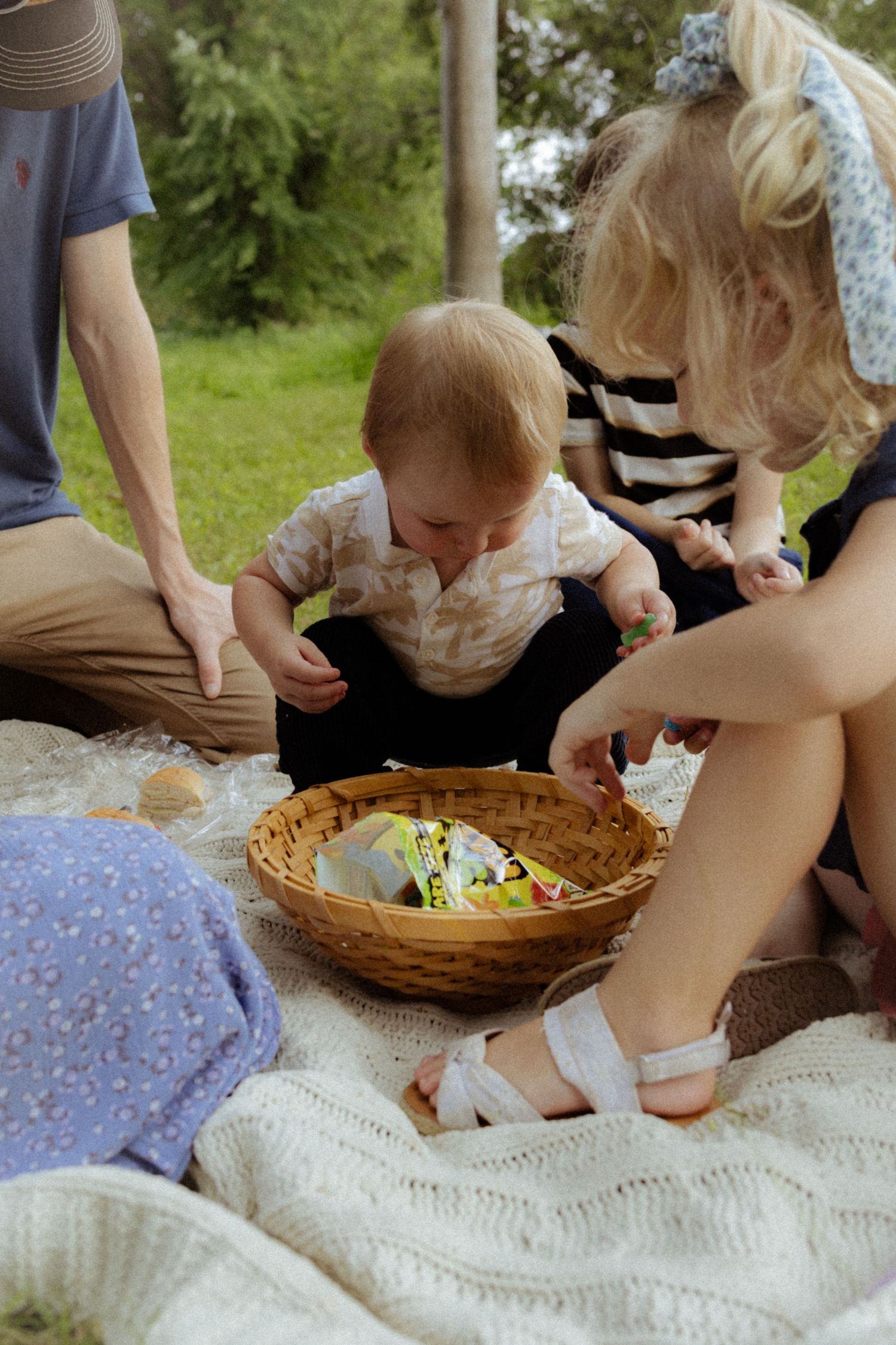Family gathered on blanket outdoors, baby exploring wicker basket with Easter eggs during spring picnic celebration.