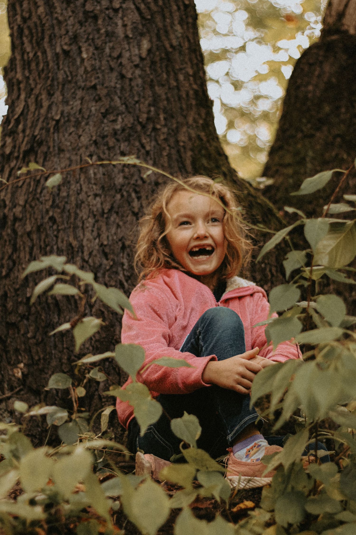 A joyful young girl with curly blonde hair sits laughing among forest foliage beside a large tree trunk.