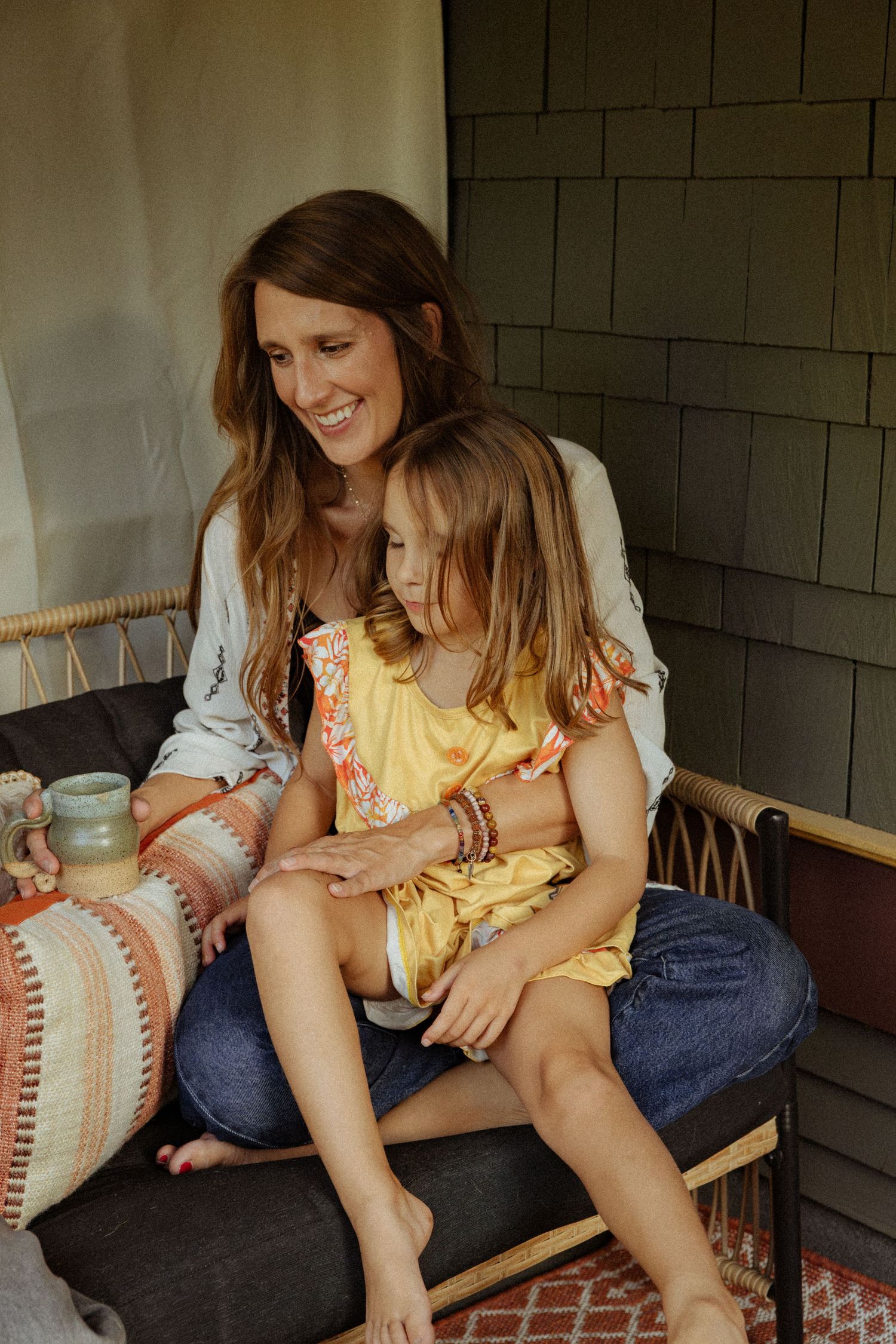 A smiling woman in a white cardigan hugs a young girl in a yellow dress on her lap on a cozy porch daybed.