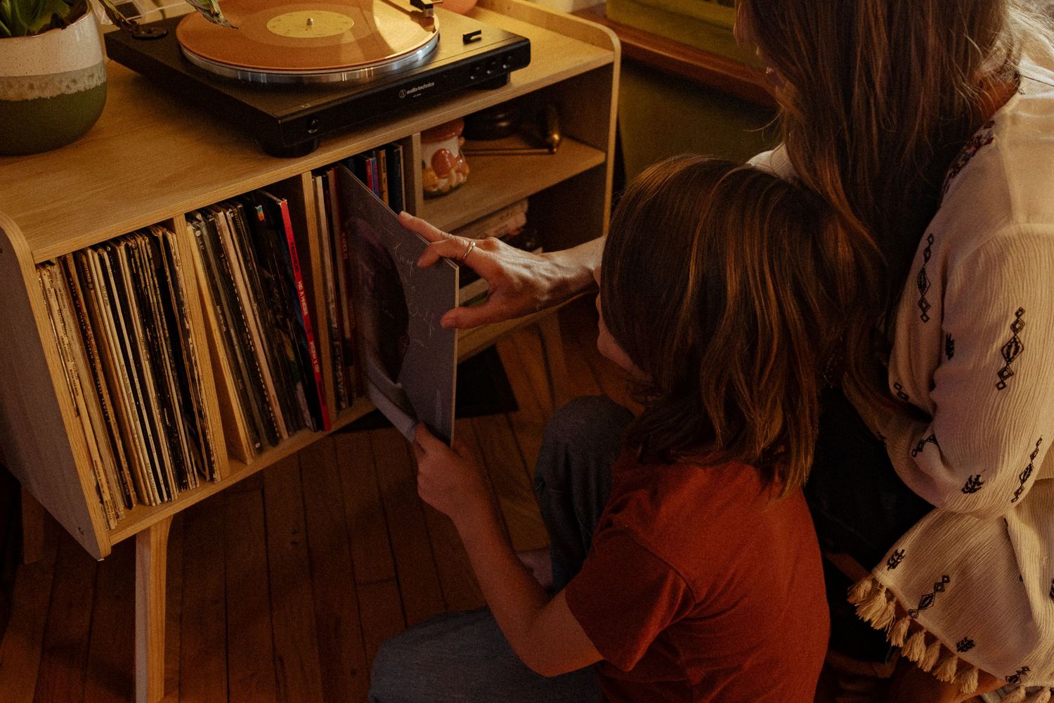 Two people crouch beside a wooden shelf of vinyl records while one pulls out an album, with a turntable playing on top.