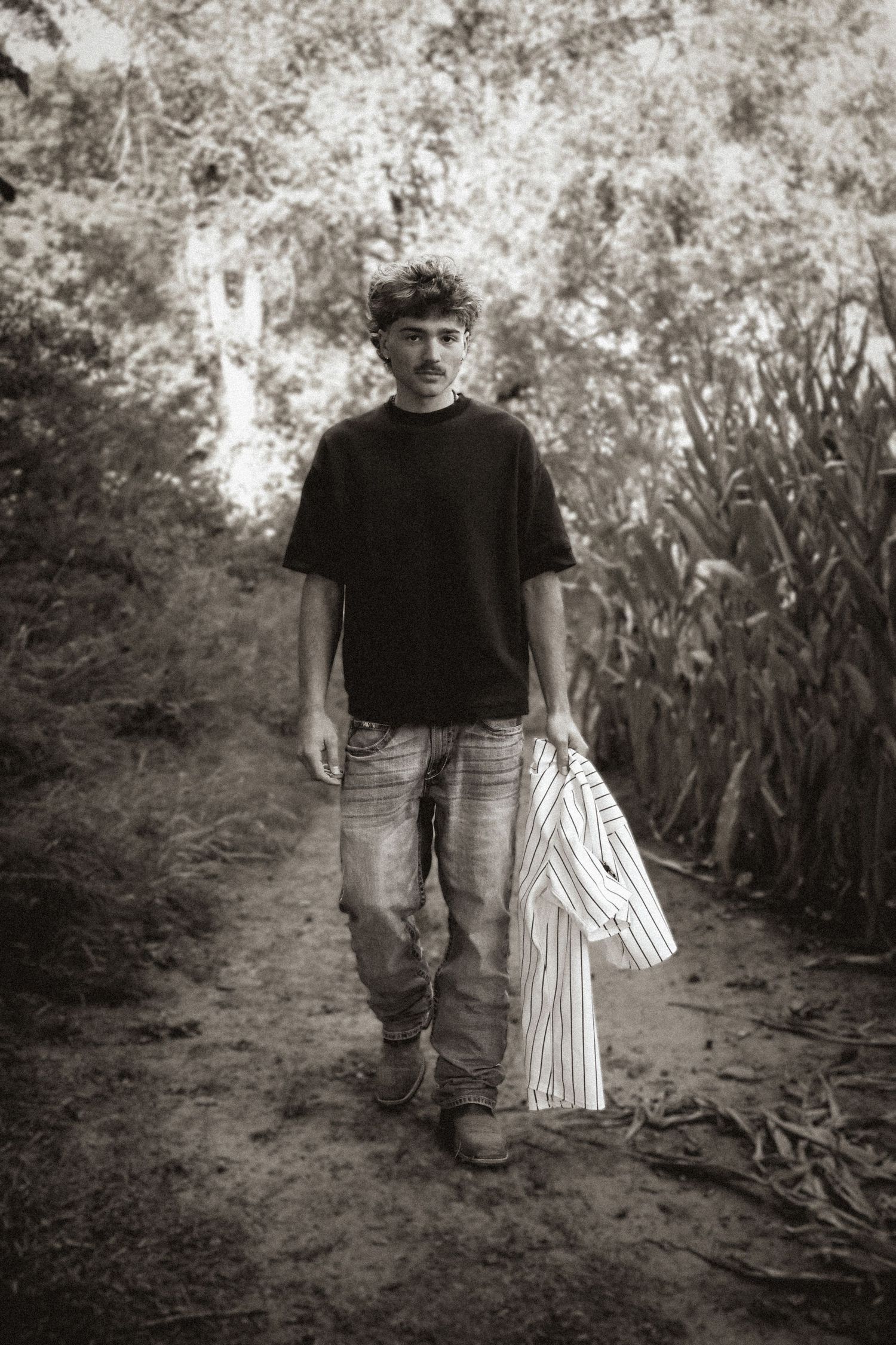 Black and white photo of a young man in jeans and black t-shirt walking on a dirt path through lush greenery.