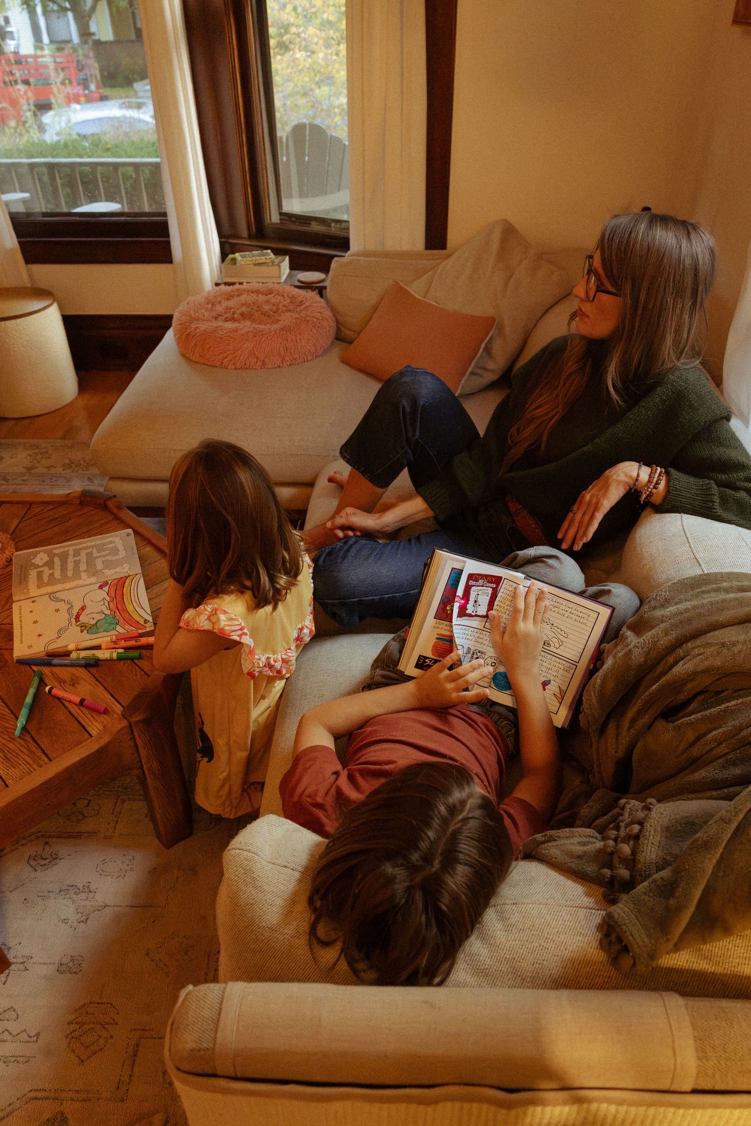 Viewed from above, a woman on a sofa smiles as one child reads a book and another colors at a wooden coffee table.