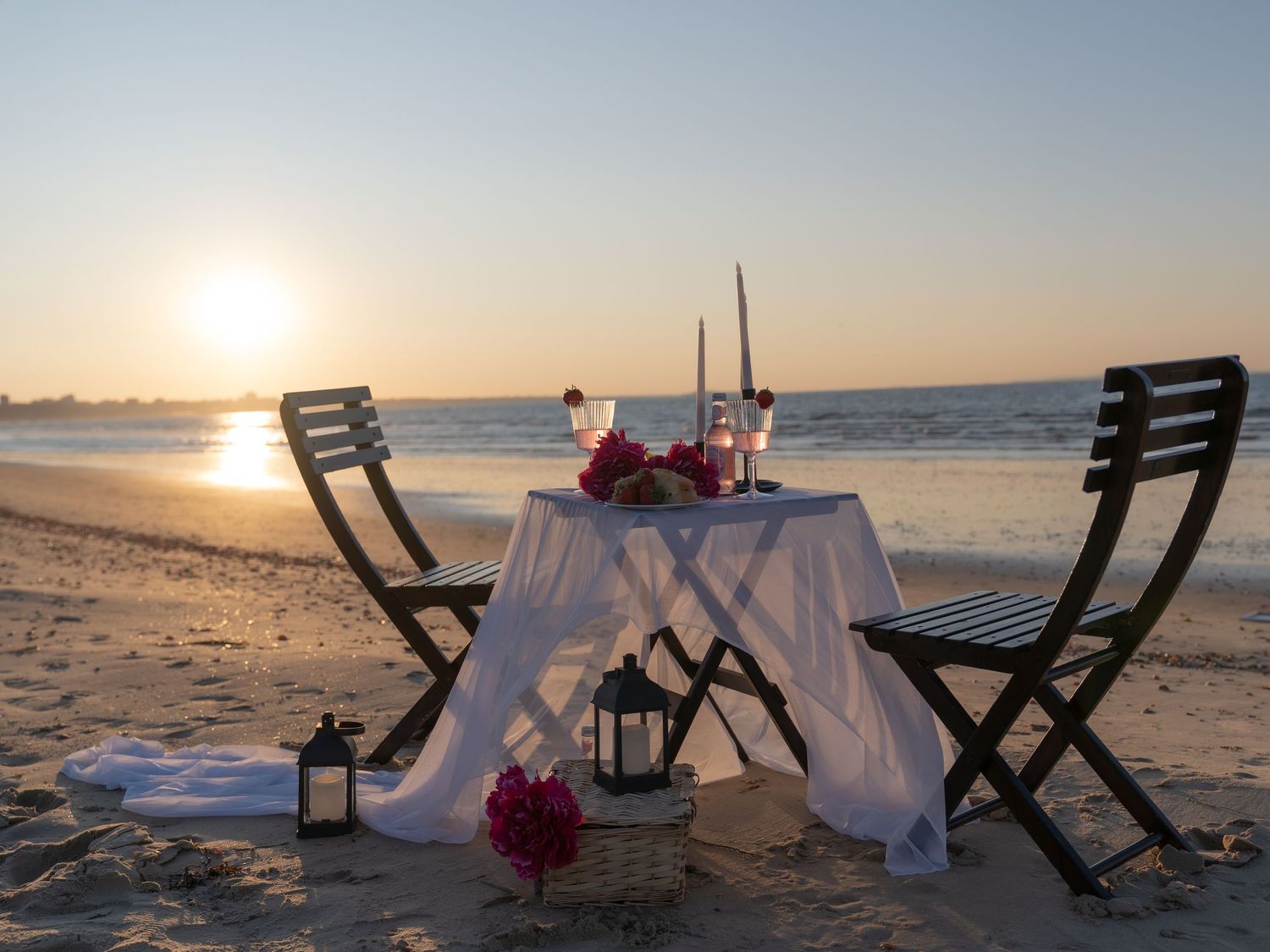 Romantic beach dinner setup for two at sunset with candles, wine glasses, and rose petals on a white draped table.