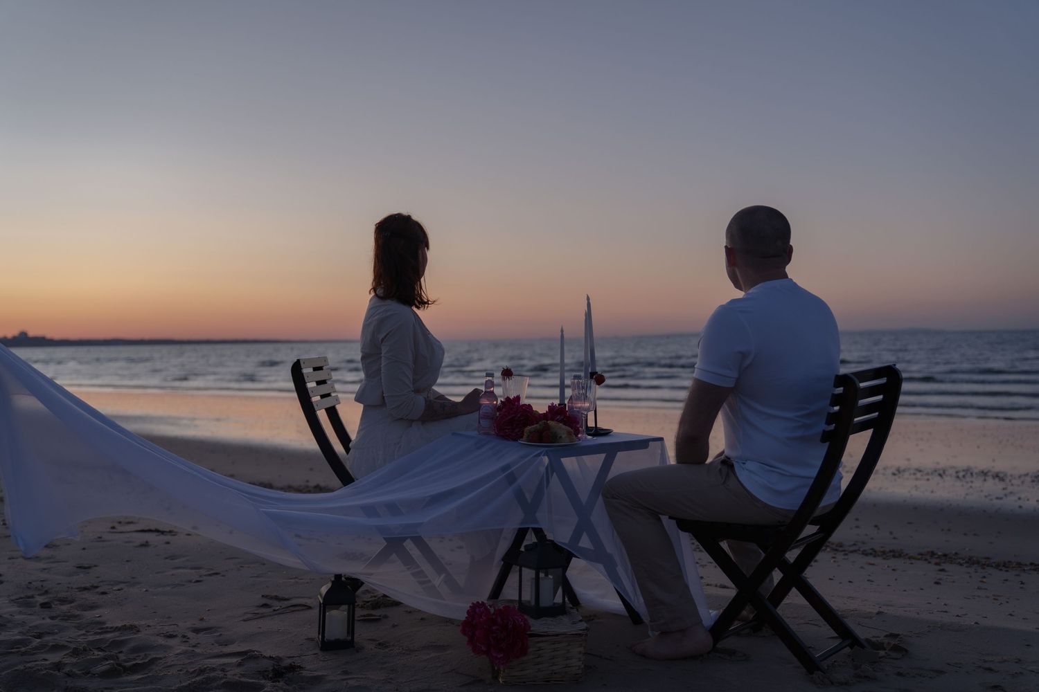 Couple enjoying a romantic candlelit dinner on a beach at sunset with ocean views.