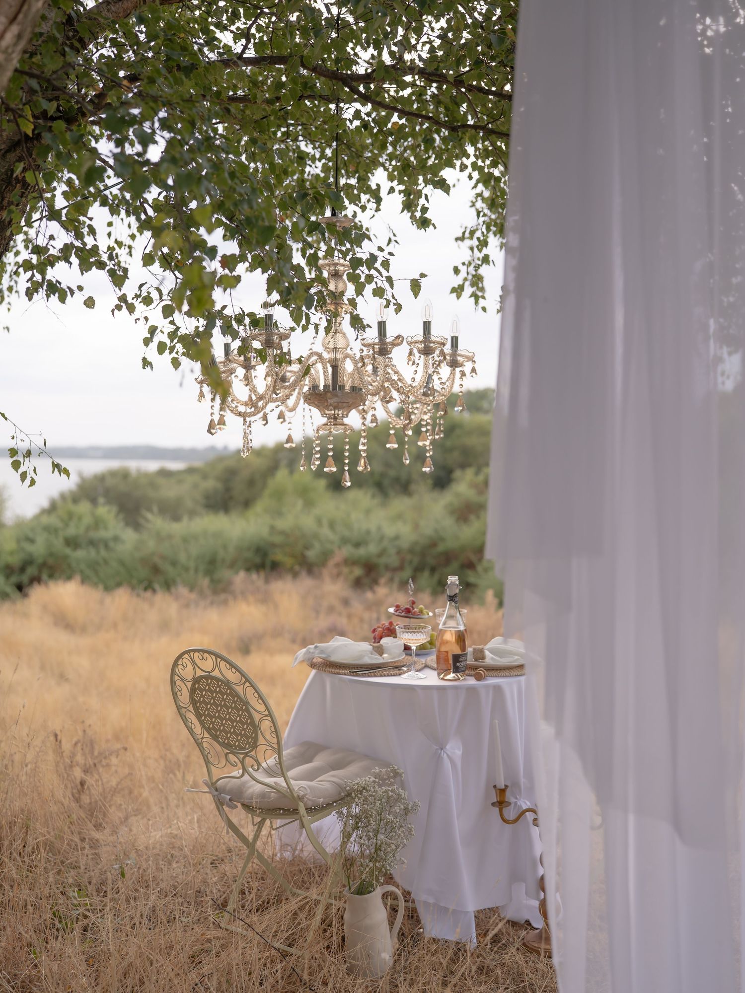 Romantic outdoor dining setup with crystal chandelier hanging from tree, white tablecloth, and sheer curtains in golden meadow.