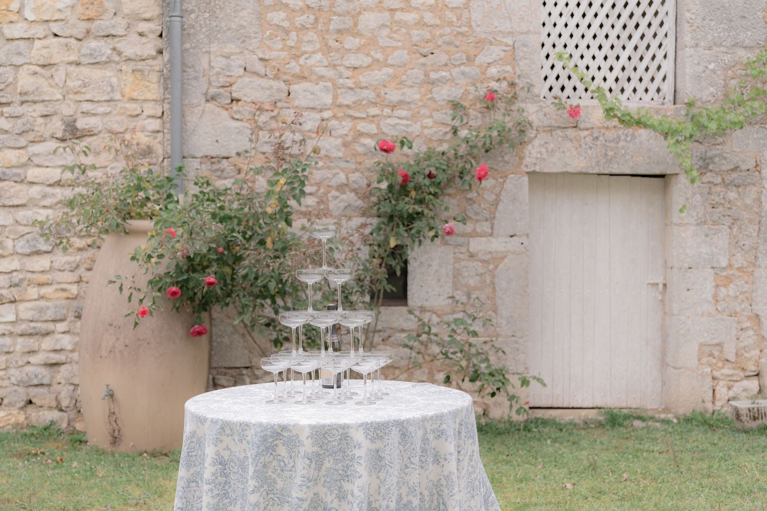 Elegant outdoor cocktail table with champagne glasses and roses climbing stone wall at a garden wedding venue.