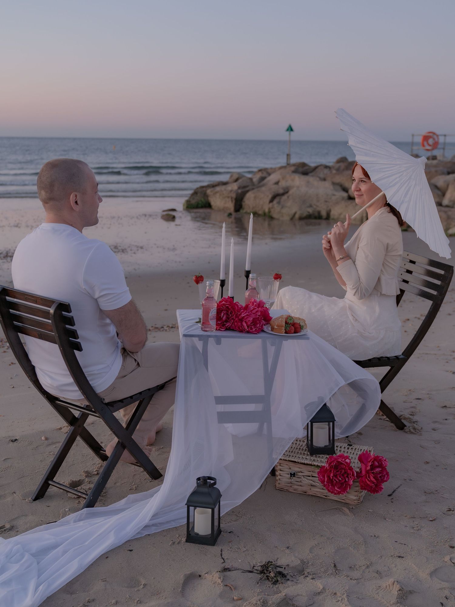 Romantic beach dinner at sunset with couple at candlelit table, pink flowers, lanterns, and white parasol by the ocean.
