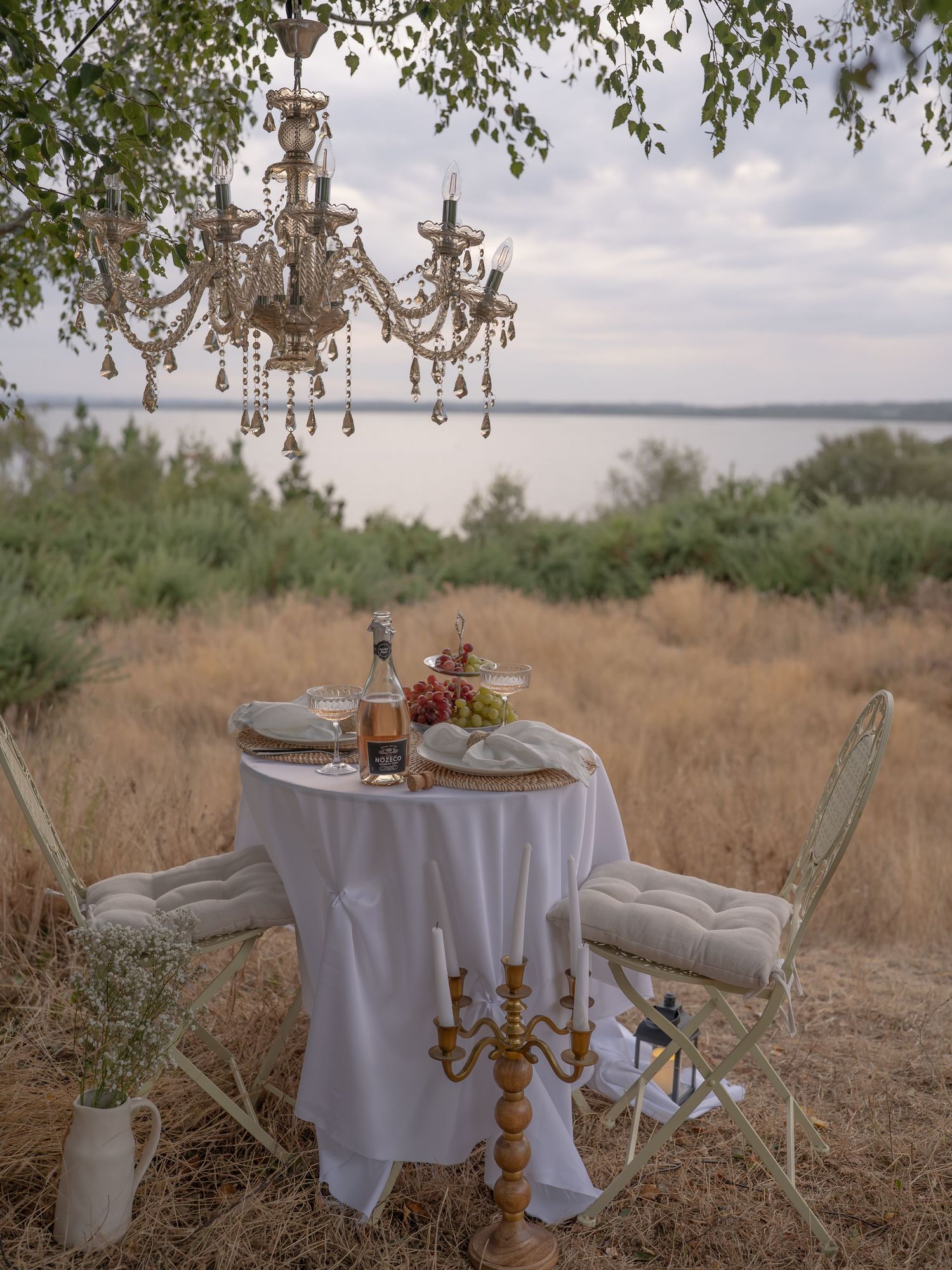 Elegant outdoor dining setup with chandeliers, white tablecloth, candelabra, and grapes near a scenic waterfront.