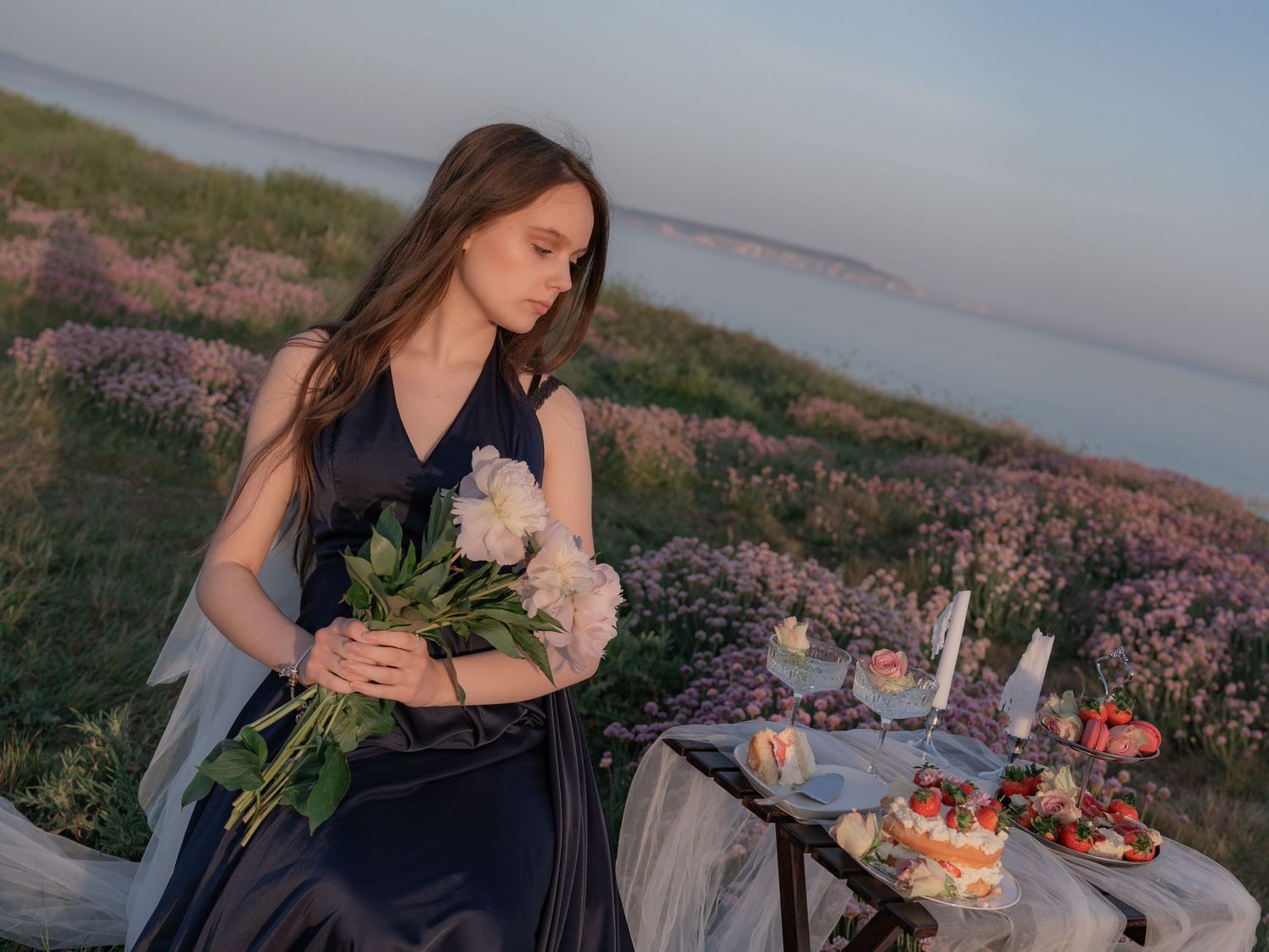 Young woman in navy dress holds white peonies at an outdoor table with desserts in a blooming field at dusk.