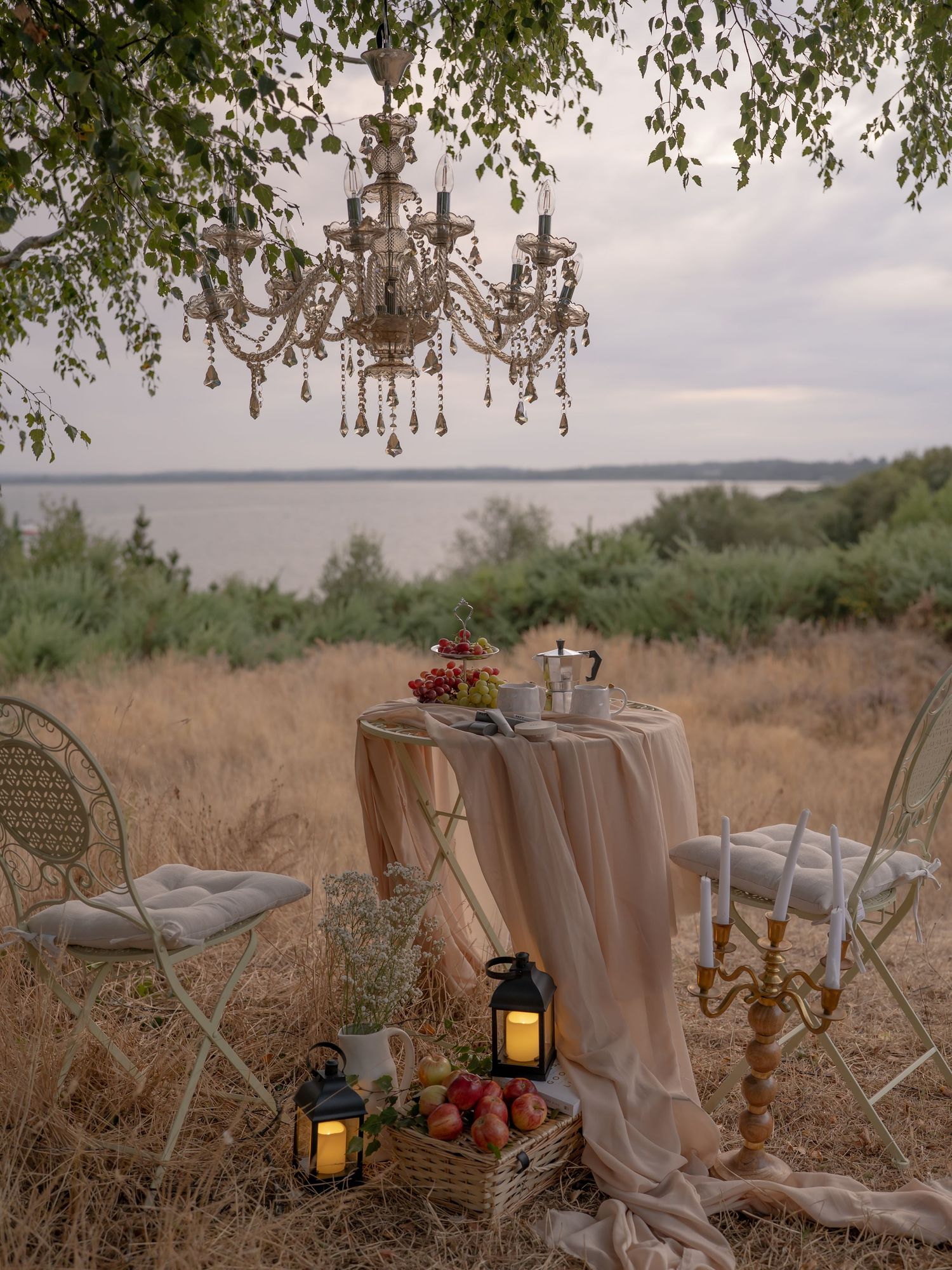 Romantic outdoor tablescape with crystal chandelier, candles, and fruit basket set in a field overlooking the sea.