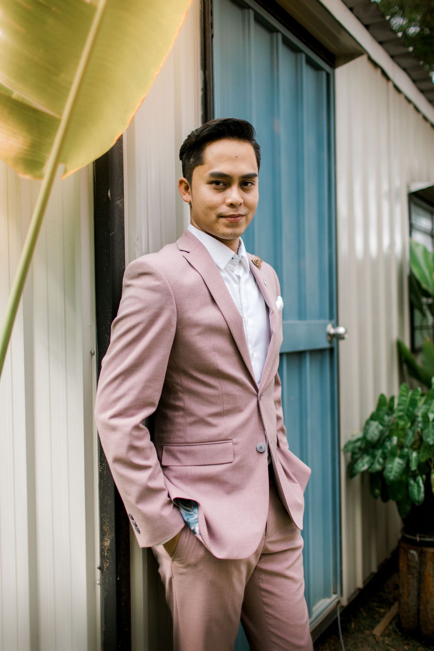 A young man in a stylish pink suit and tie poses confidently outdoors near a blue door and tropical plants.
