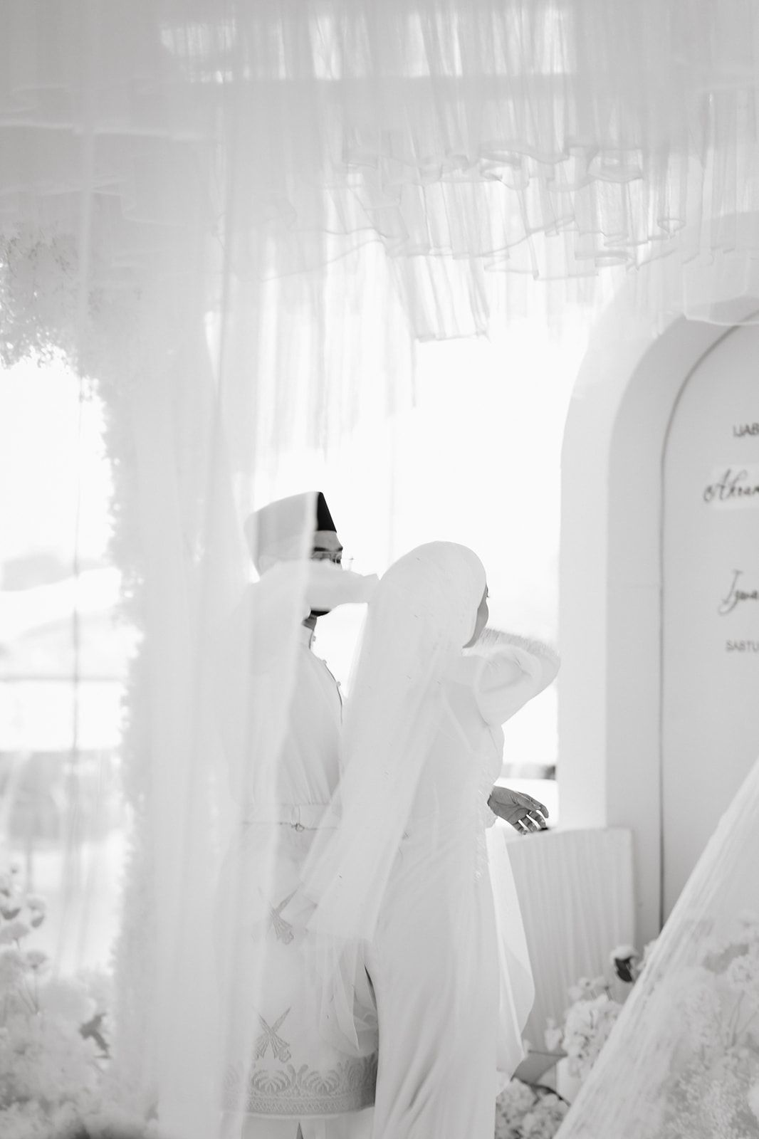 Black and white photo of MuMalaysian lim bride adjusting her hijab at a white floral wedding venue.