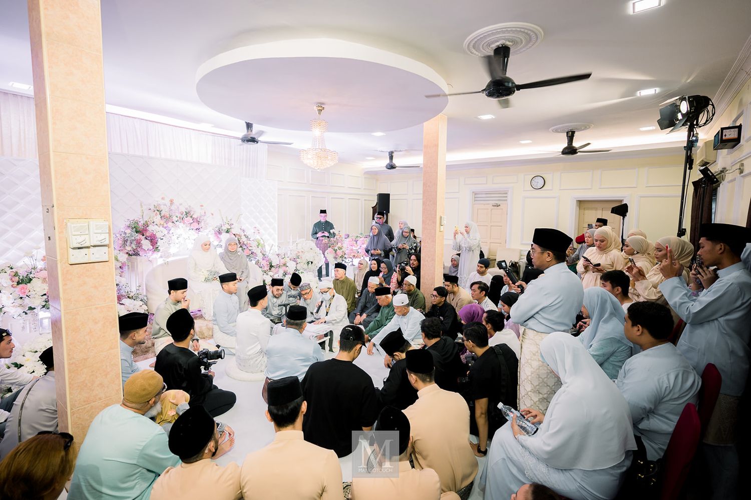 Wide view of a crowded Malaysian wedding ceremony held indoors with floral decorations in the background.