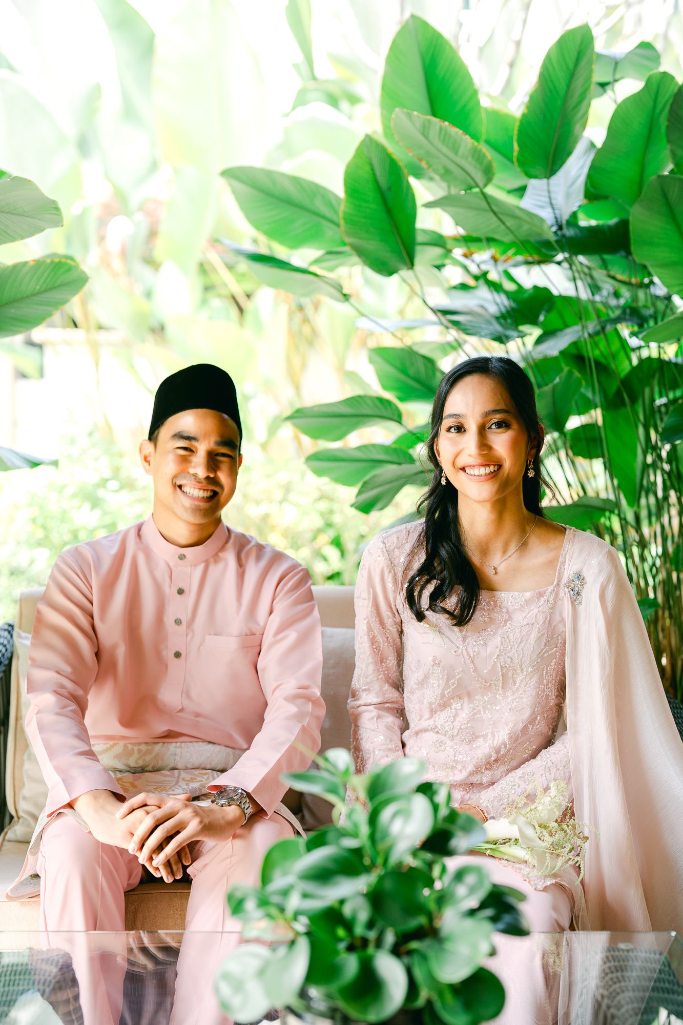 Malay couple in matching pink traditional baju kurung and baju melayu sitting together surrounded by tropical greenery.