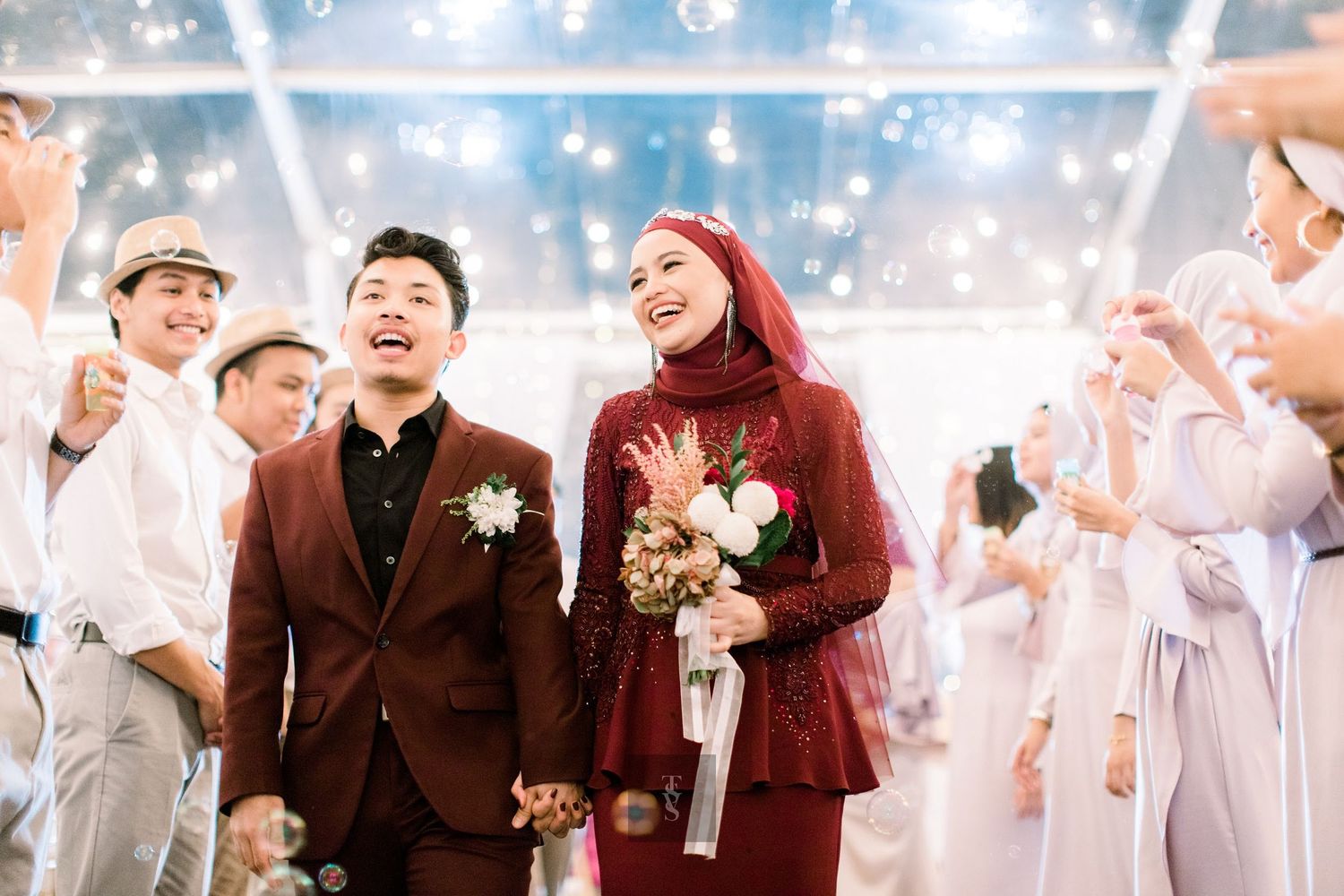 A joyful couple in matching burgundy wedding attire walks through a festive celebration with bubbles and guests.