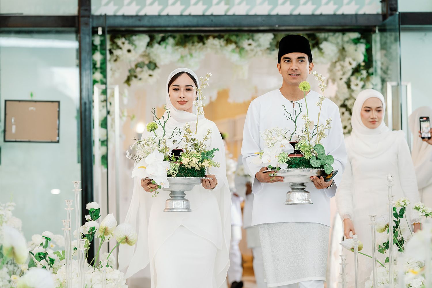 Malaysian couple in white traditional attire carrying floral centerpieces at wedding reception. Featuring Syed Saddiq as groomsmen
