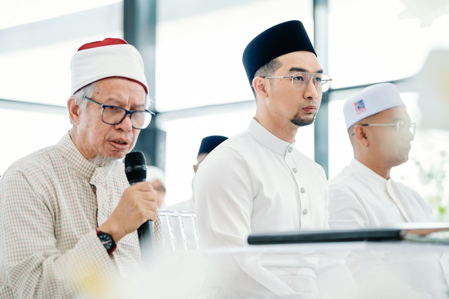 Malaysian officiant speaking at Islamic wedding ceremony with groom seated beside him.