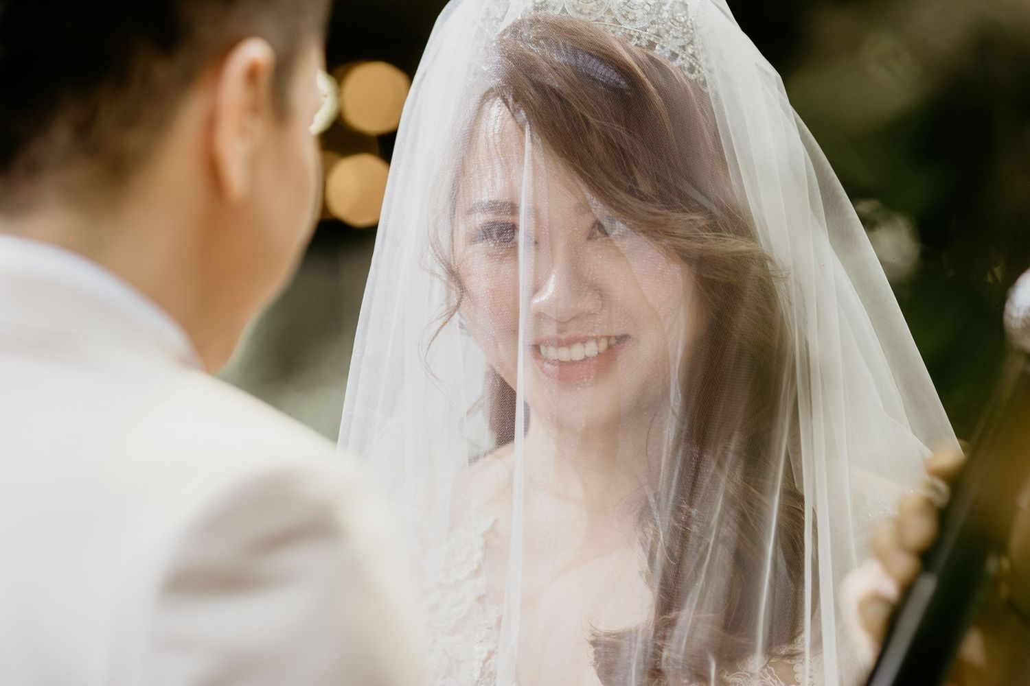 Radiant bride smiling through sheer veil at groom during emotional outdoor wedding ceremony.