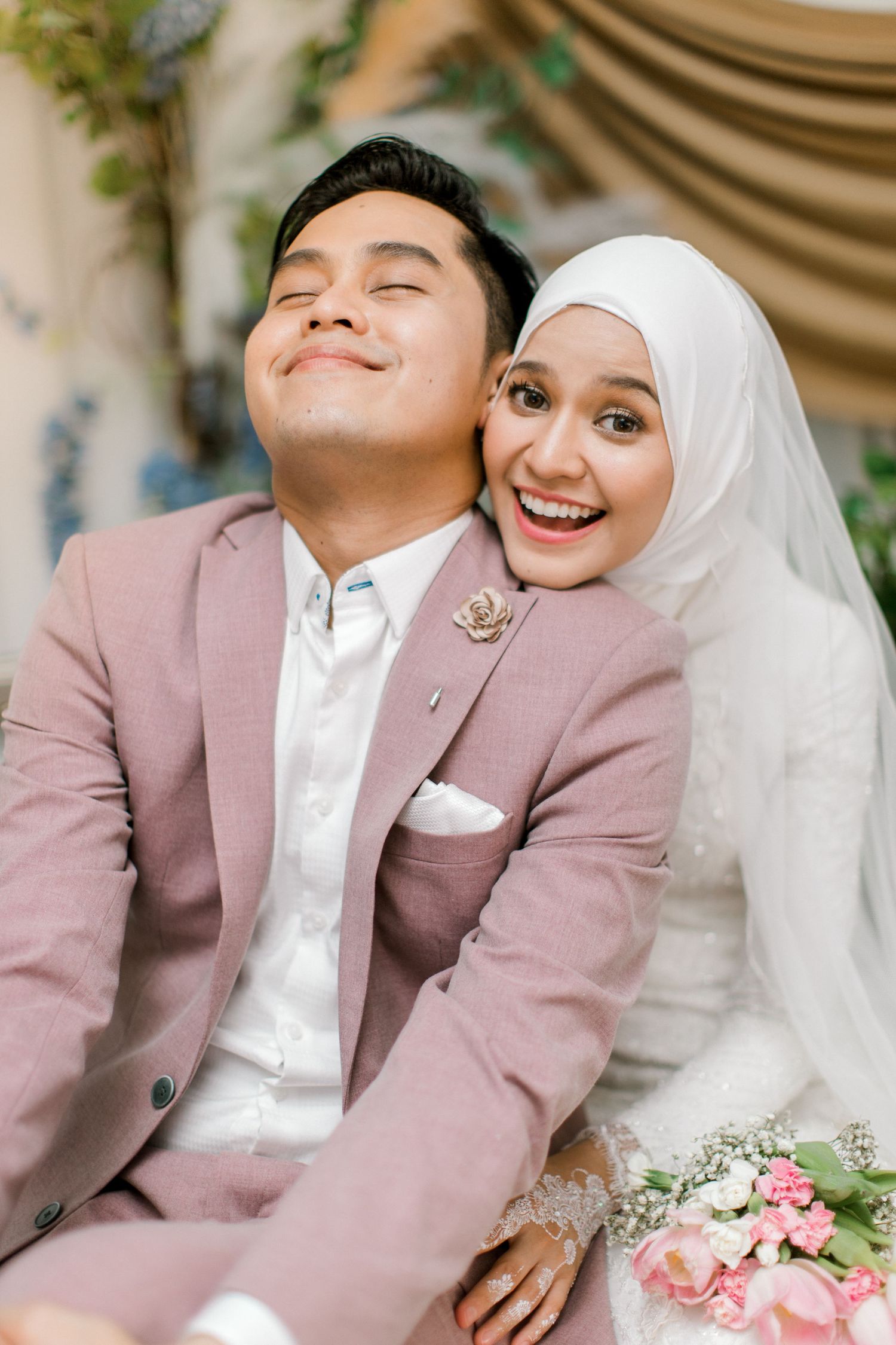 Malaysian bride in white hijab and groom in pink suit smiling together at their wedding ceremony.