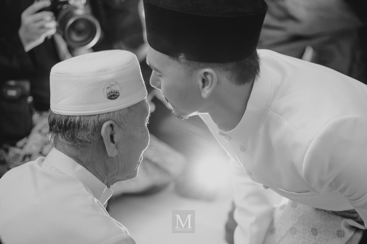 Black and white photo of an elder and young man in traditional Malaysian attire sharing an intimate moment.