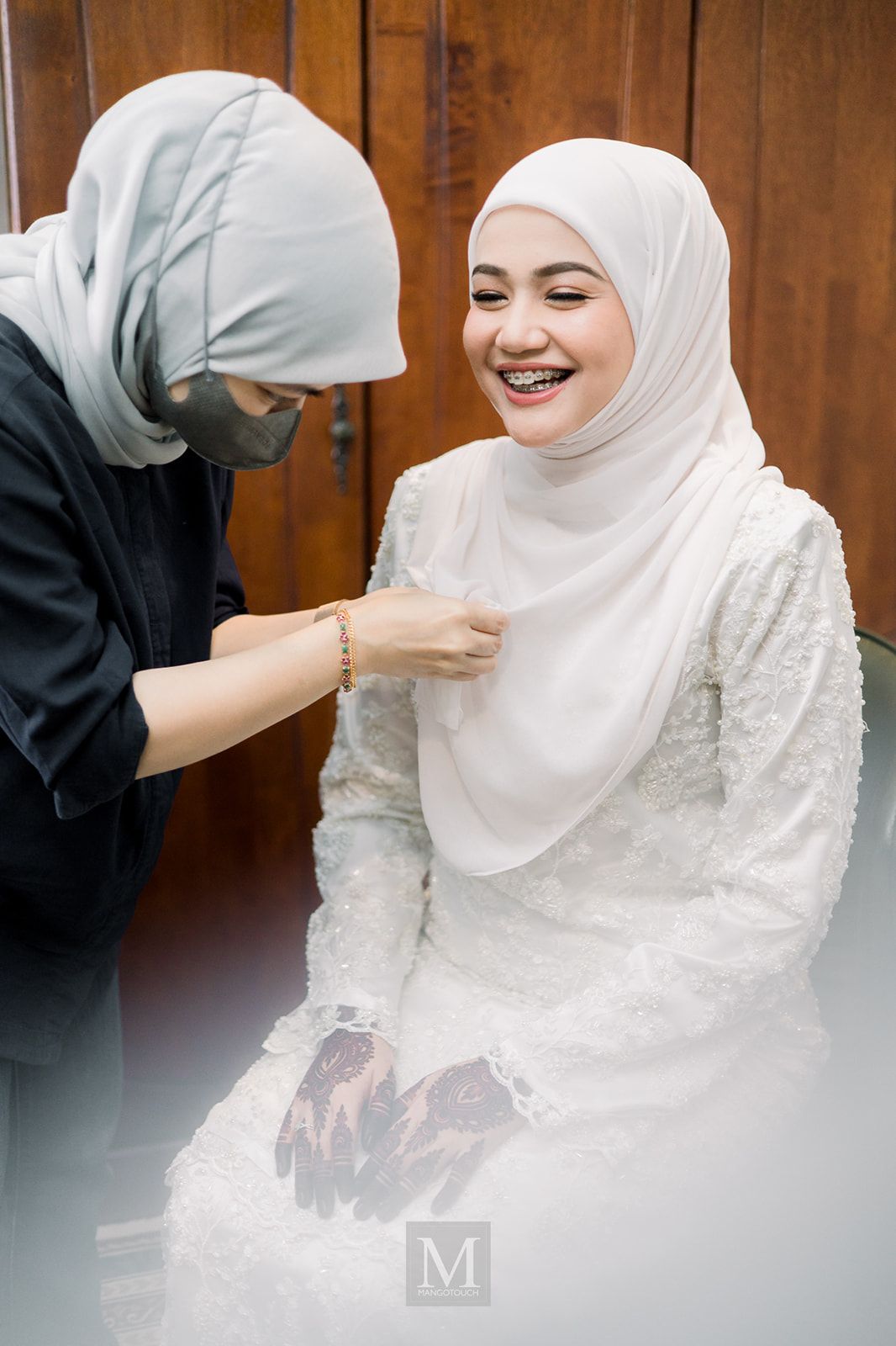 Malaysian bride in white hijab laughing joyfully while being assisted with her wedding dress.