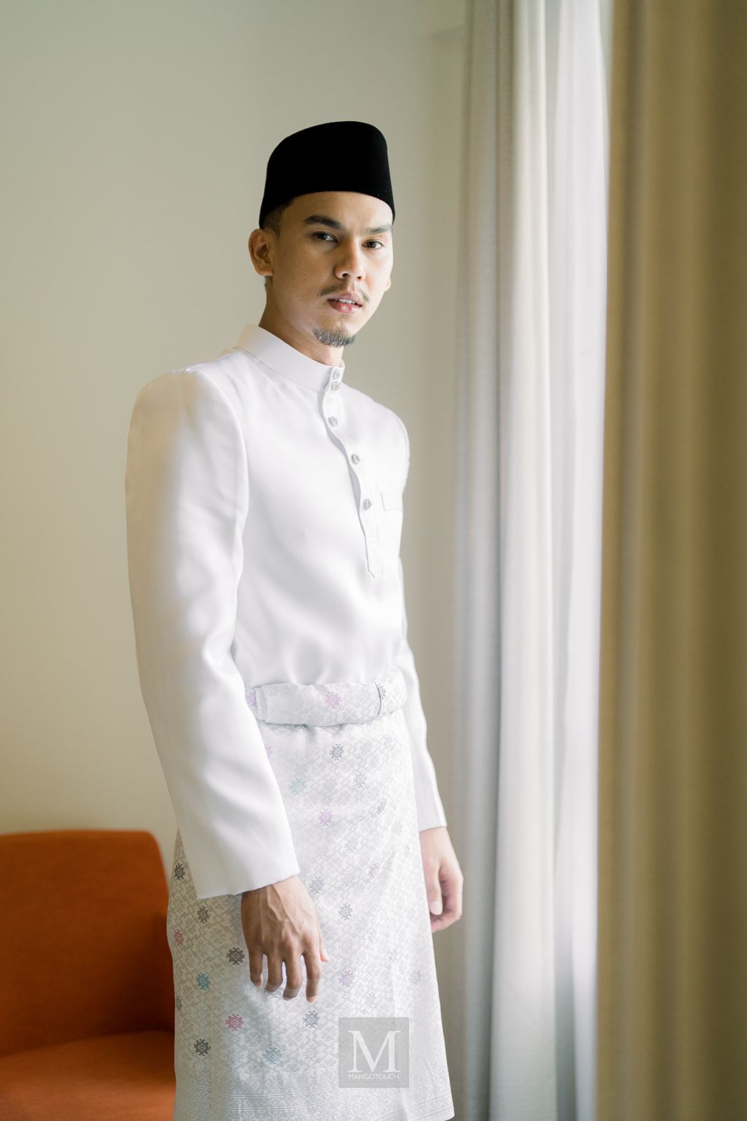Malaysian groom in traditional white baju melayu and black songkok standing by a curtained window.