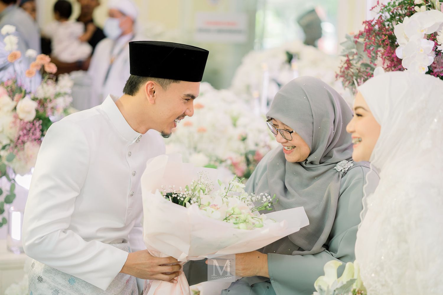Malaysian groom offering white flowers to a woman in grey hijab during a traditional Malay wedding ceremony.