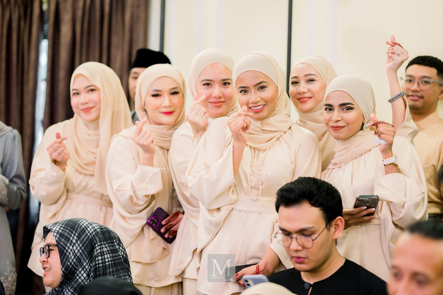 Group of women in matching cream hijabs and outfits smiling and posing together at a Malaysian wedding celebration.