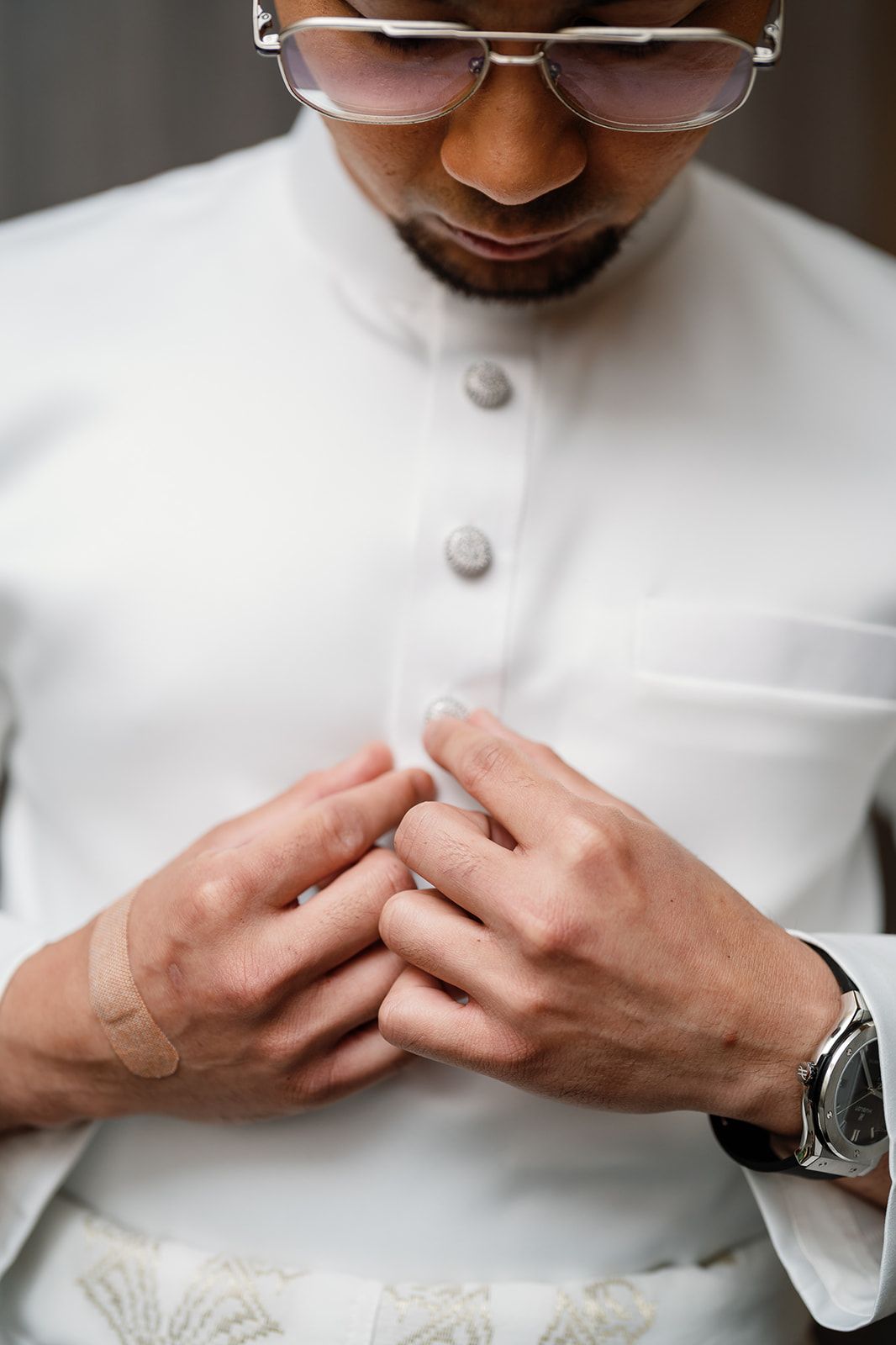 Groom in white traditional attire adjusts his buttons while wearing sunglasses and a luxury watch.