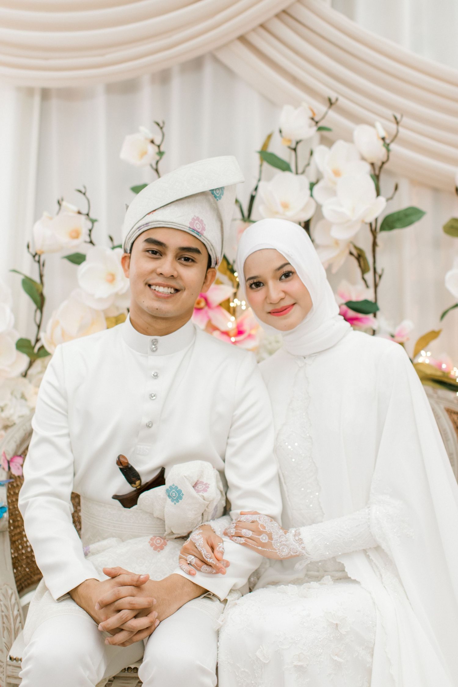 Malaysian  newlyweds in white traditional attire smiling together at floral wedding ceremony backdrop.