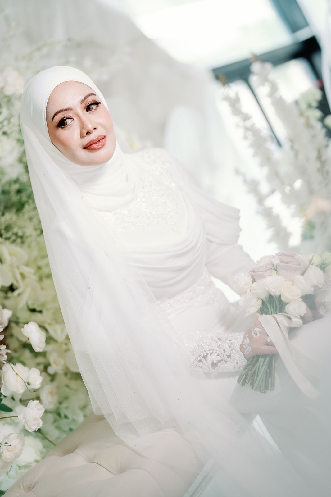 Elegant Malaysian bride in white hijab and lace gown holding a bouquet surrounded by white flowers.