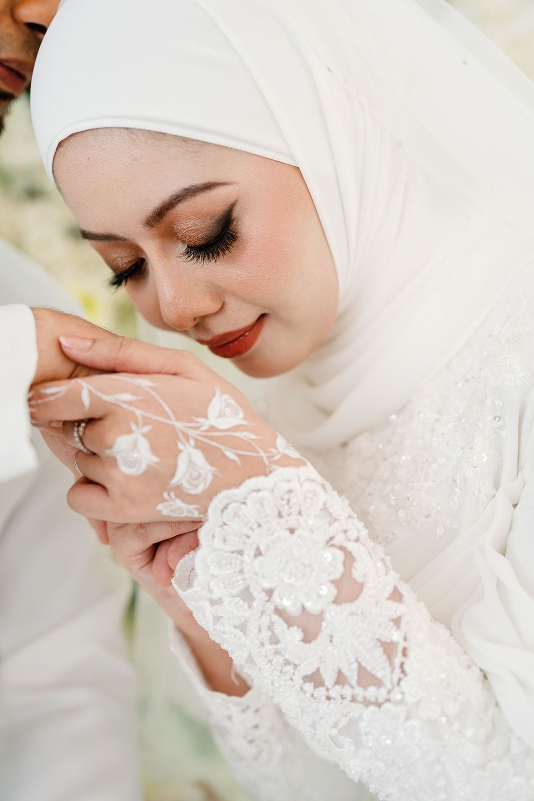 Beautiful Malaysian bride in white hijab and lace dress holding groom's hands at wedding ceremony.