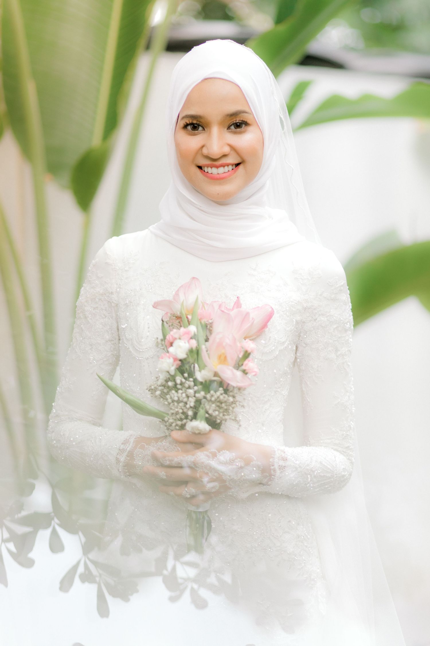 Smiling Malaysian bride in white hijab and lace gown holding pink tulip bouquet, standing among green plants.
