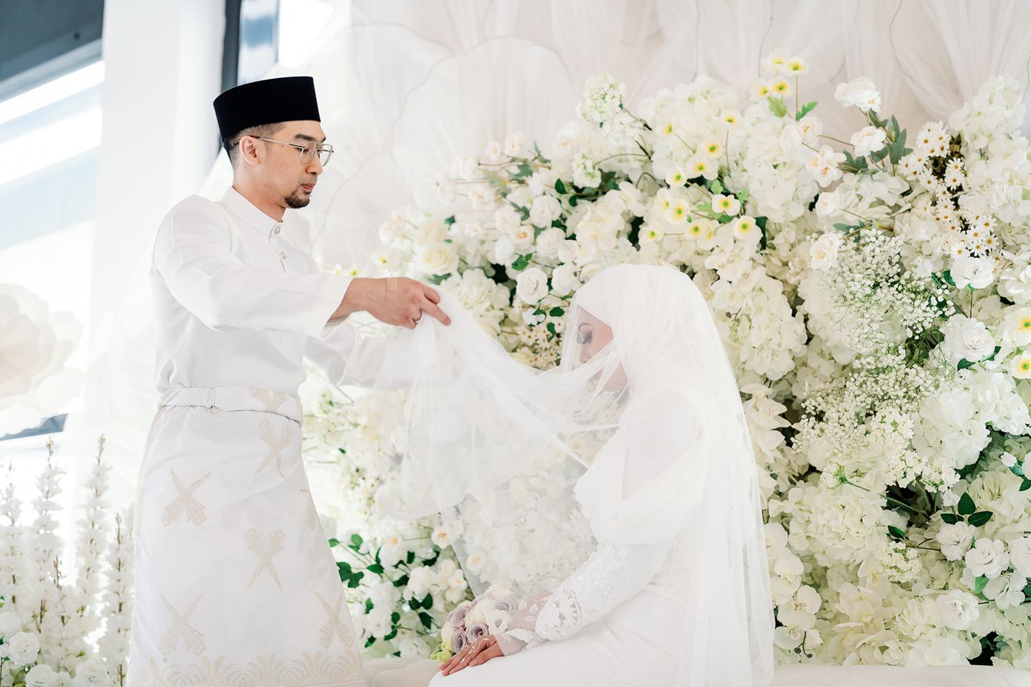 Malaysian groom lifting bride's veil beside white floral backdrop at wedding ceremony.