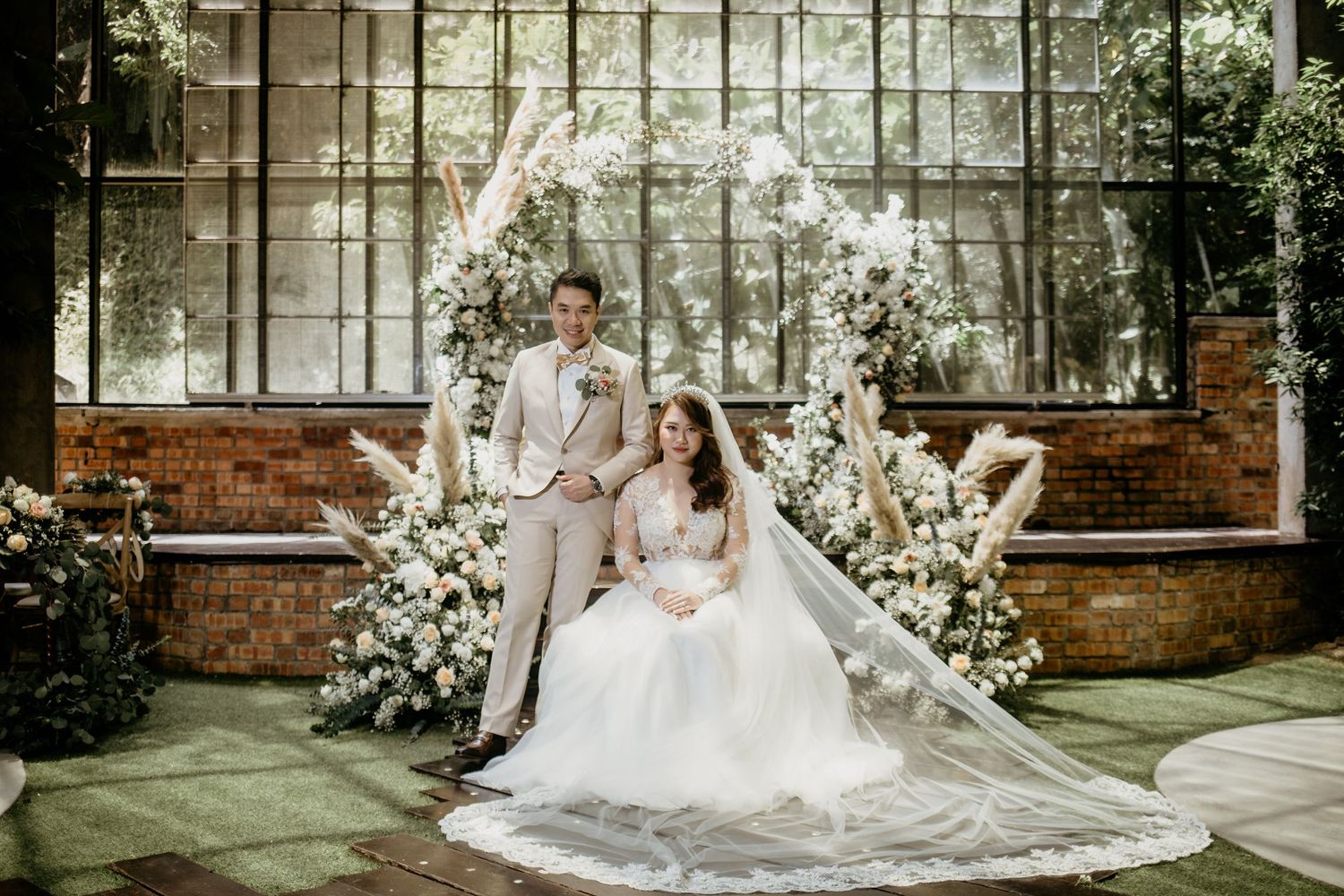 Bride and groom pose elegantly in front of a white floral arch inside a rustic greenhouse wedding venue.