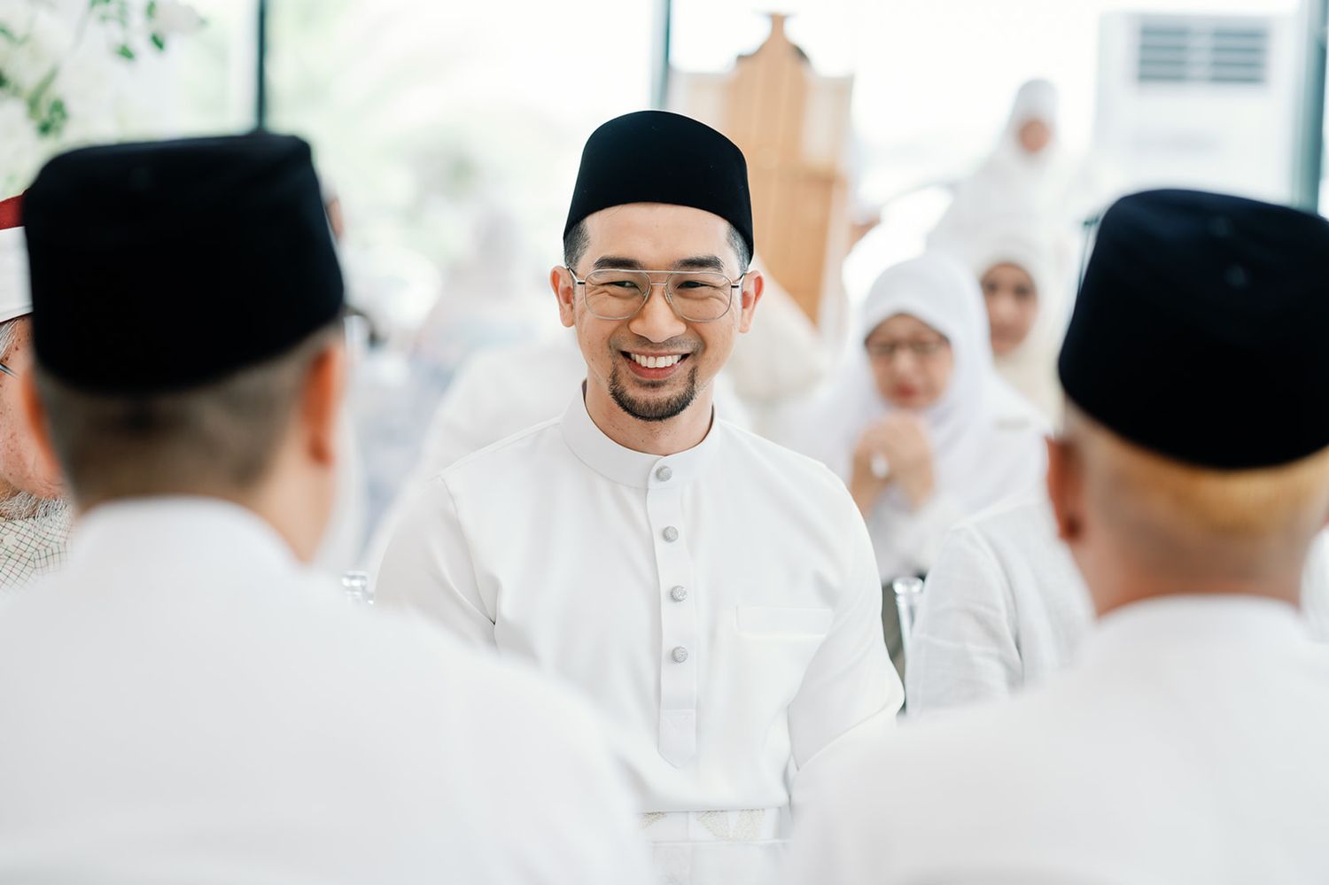 Smiling Malaysian groom in white baju melayu greeting guests at his wedding ceremony.