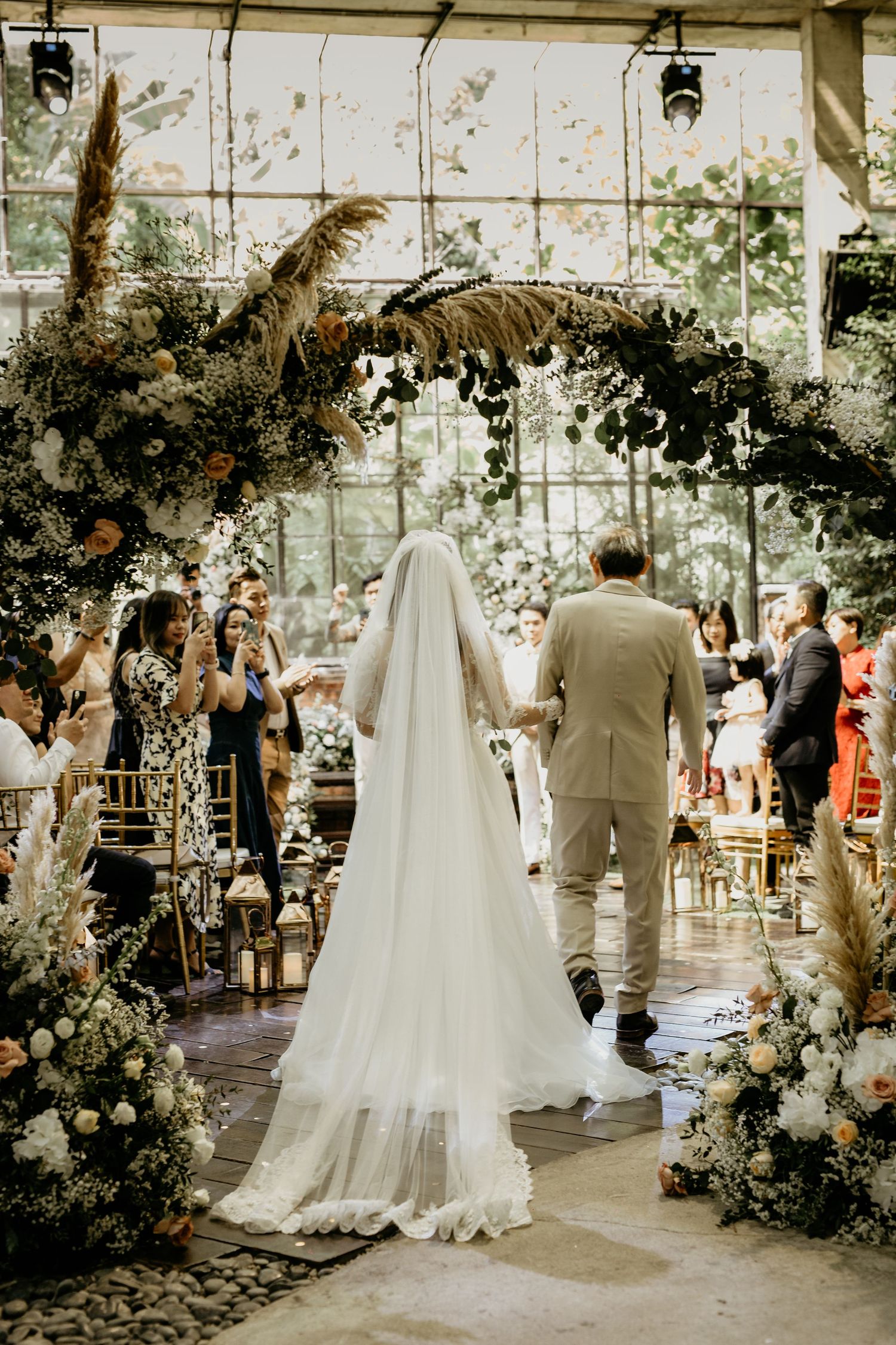 Bride in white veil and gown walks down aisle under lush floral arch with wedding guests seated.