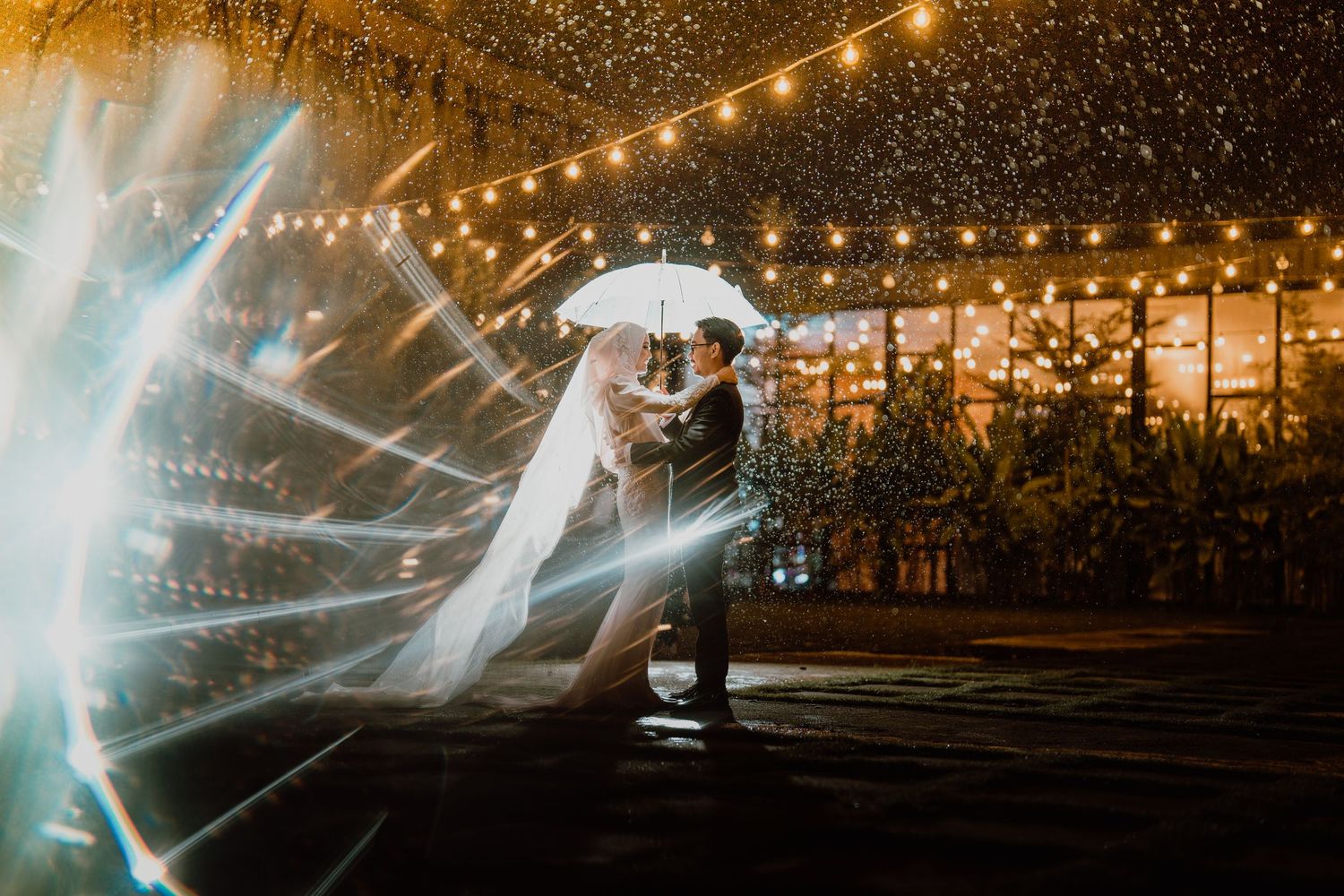 Bride and groom share a romantic dance under an umbrella in the rain with festive string lights.