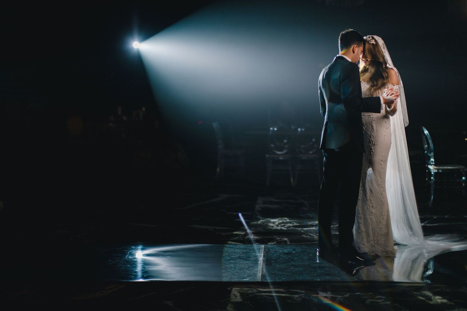 Bride and groom sharing a romantic first dance kiss under a dramatic spotlight on a dark stage.