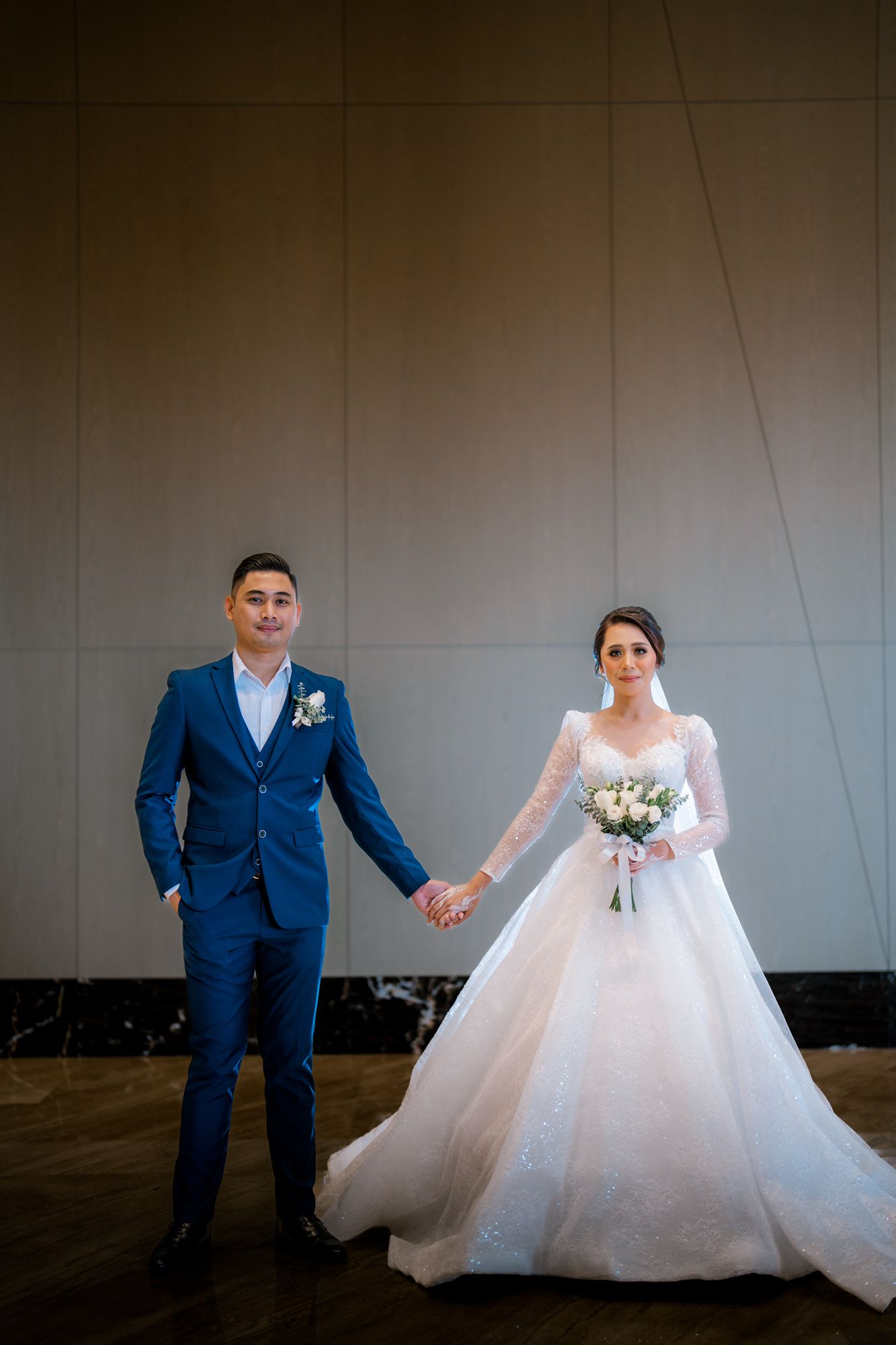 Newlywed couple holding hands, groom in blue suit and bride in elegant white ball gown with bouquet.