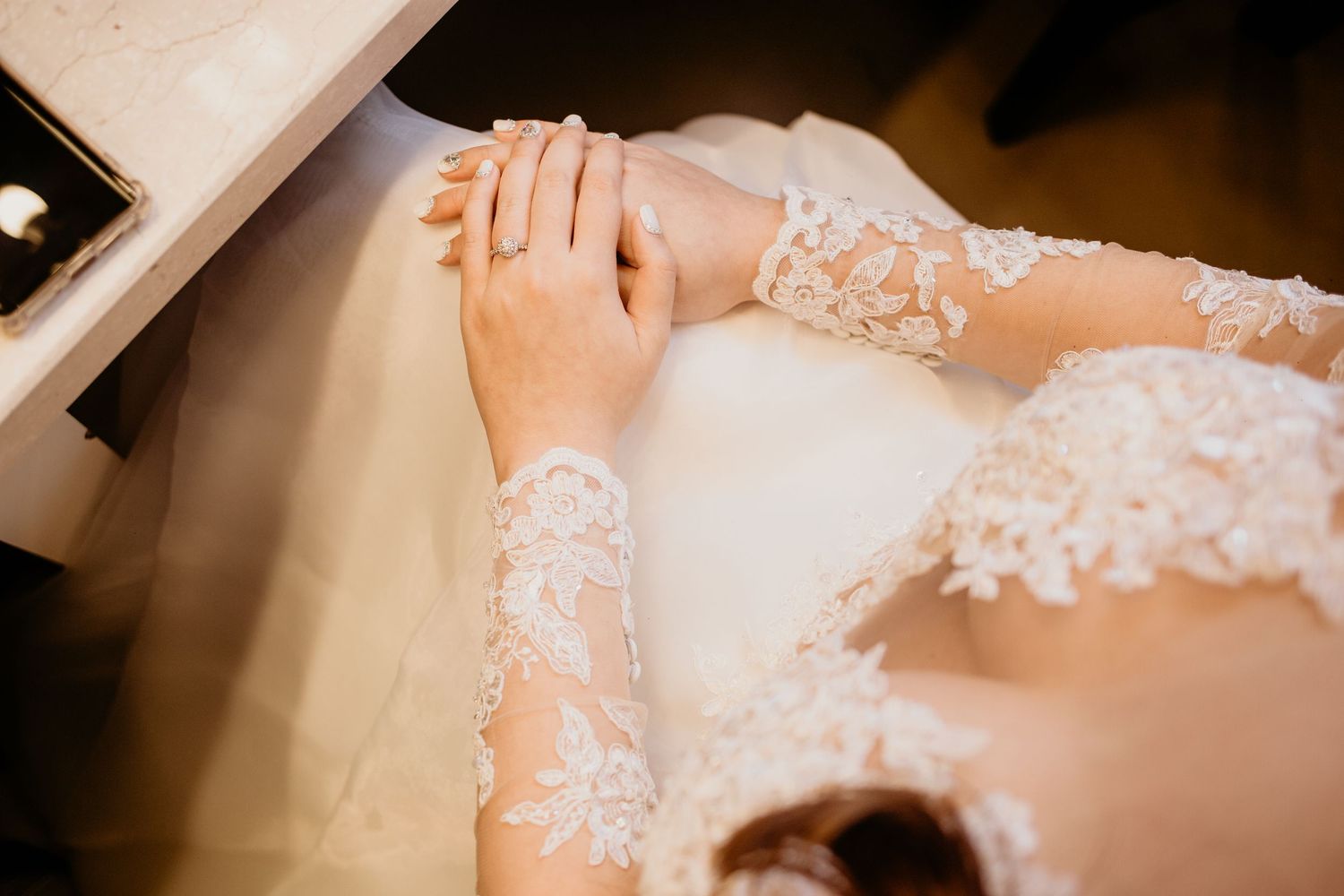 Close-up of bride's hands resting on lace wedding dress with intricate floral embroidery details.