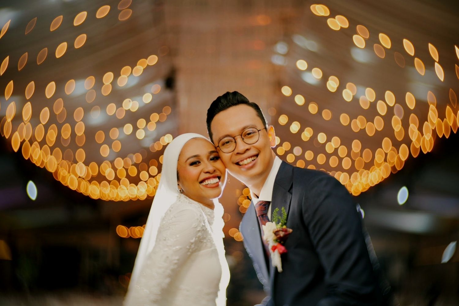 Smiling newlywed couple posing under warm bokeh lights, bride in hijab and groom in suit.