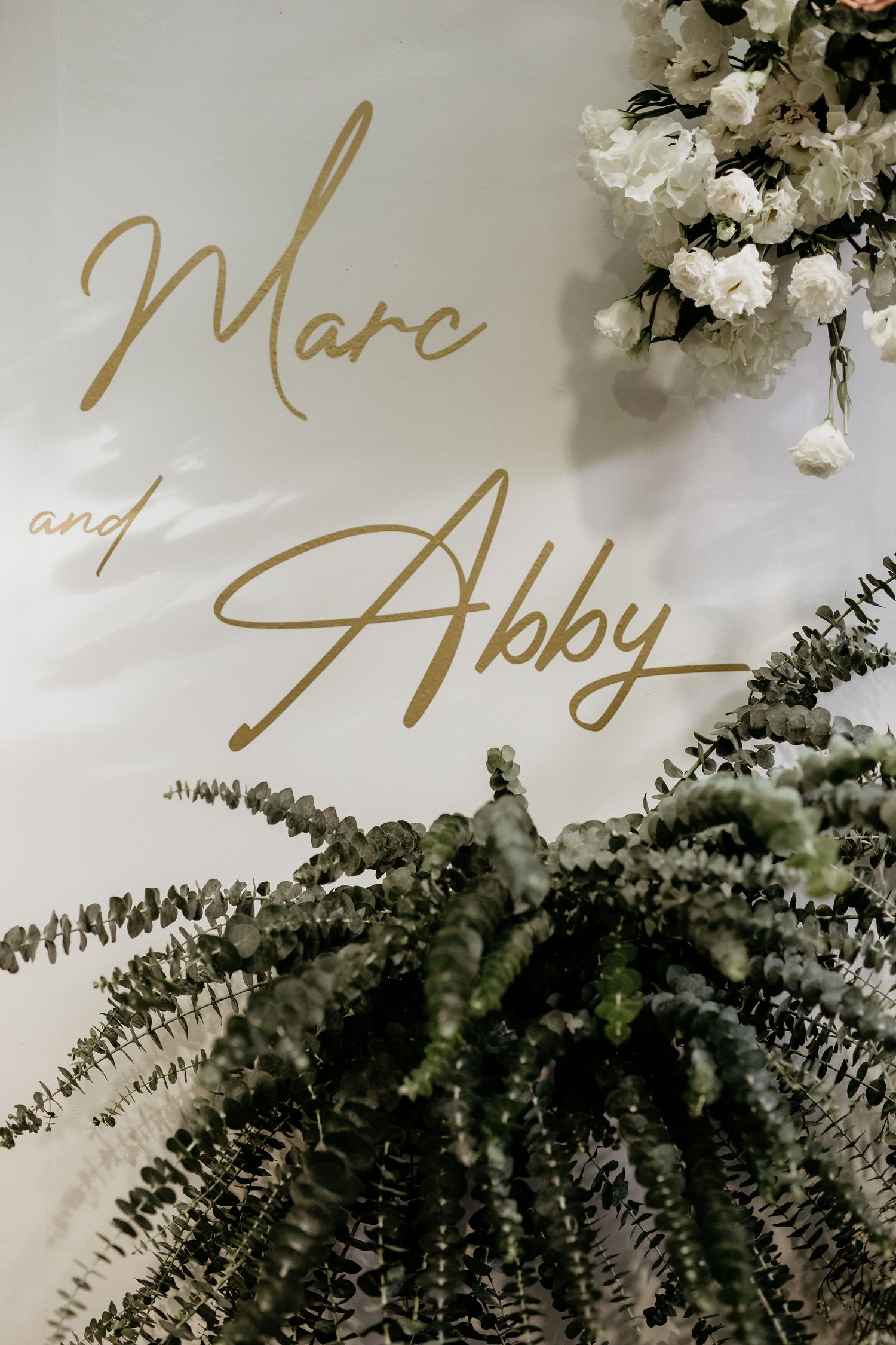 Gold script Marc and Abby wedding backdrop sign adorned with white flowers and lush green ferns.