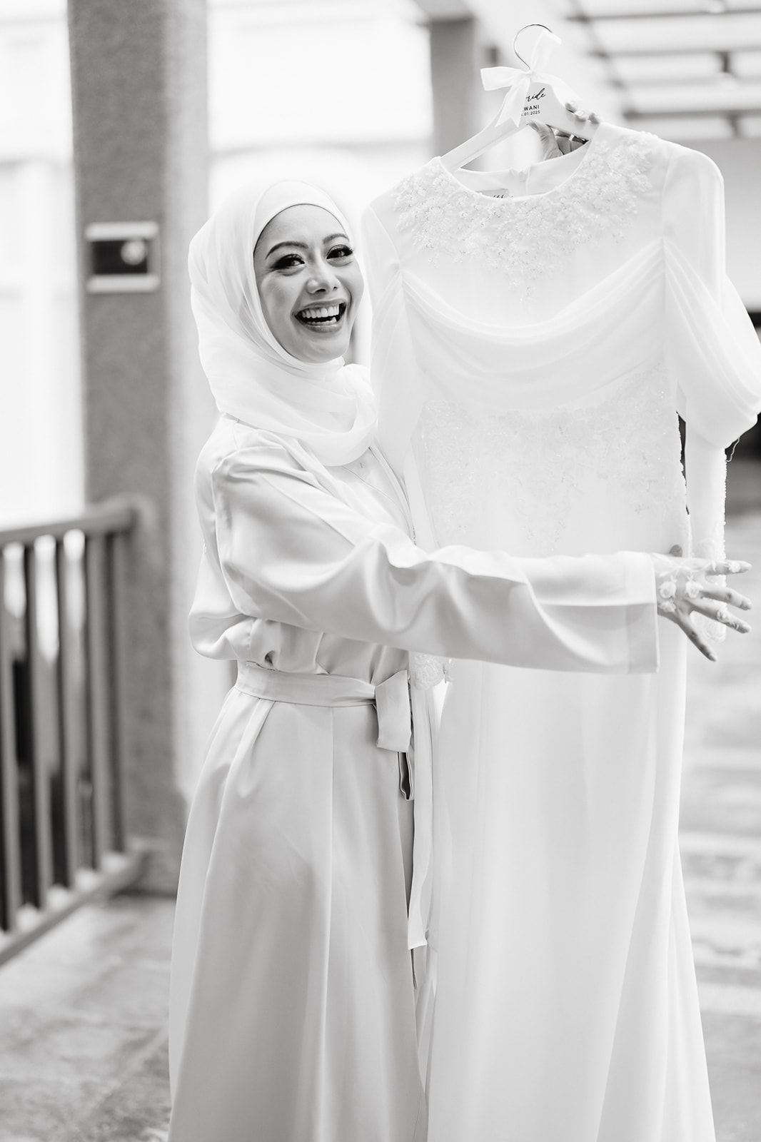 Laughing bride in hijab holds up her white wedding dress on a hanger in black and white photo.