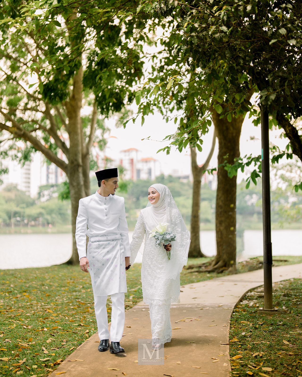 Malaysian couple in white wedding attire holding hands walking along tree-lined park pathway.