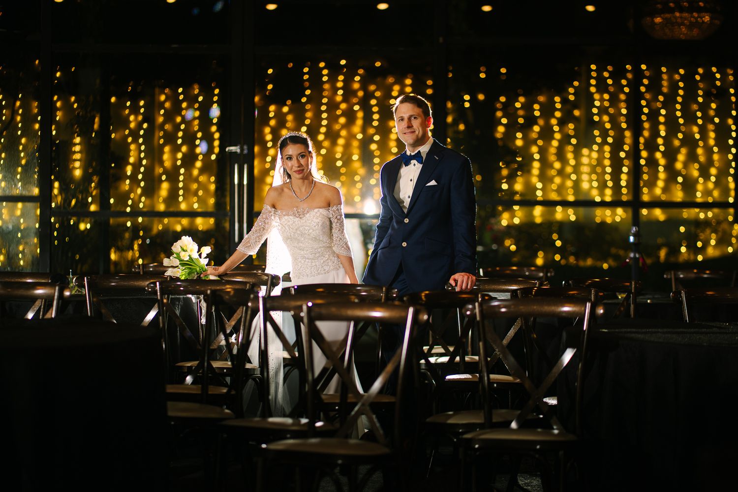 Bride and groom standing elegantly among reception tables adorned with warm fairy light backdrop.