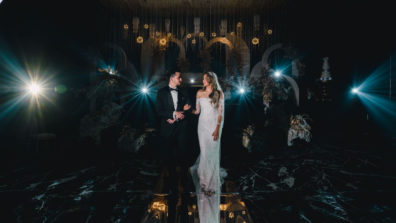 Bride and groom sharing a romantic moment on a dark reflective floor with glowing stage lights.