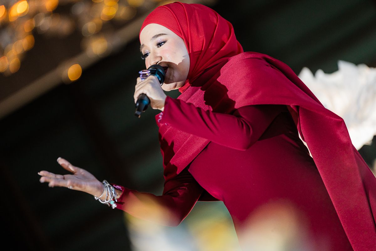 Female performer in red hijab and outfit singing passionately into microphone on stage.