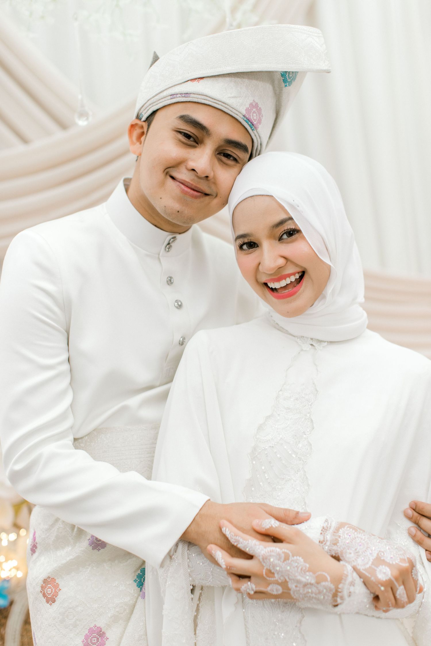 Smiling Malaysian couple in white traditional Malay wedding attire embracing at decorated ceremony venue.